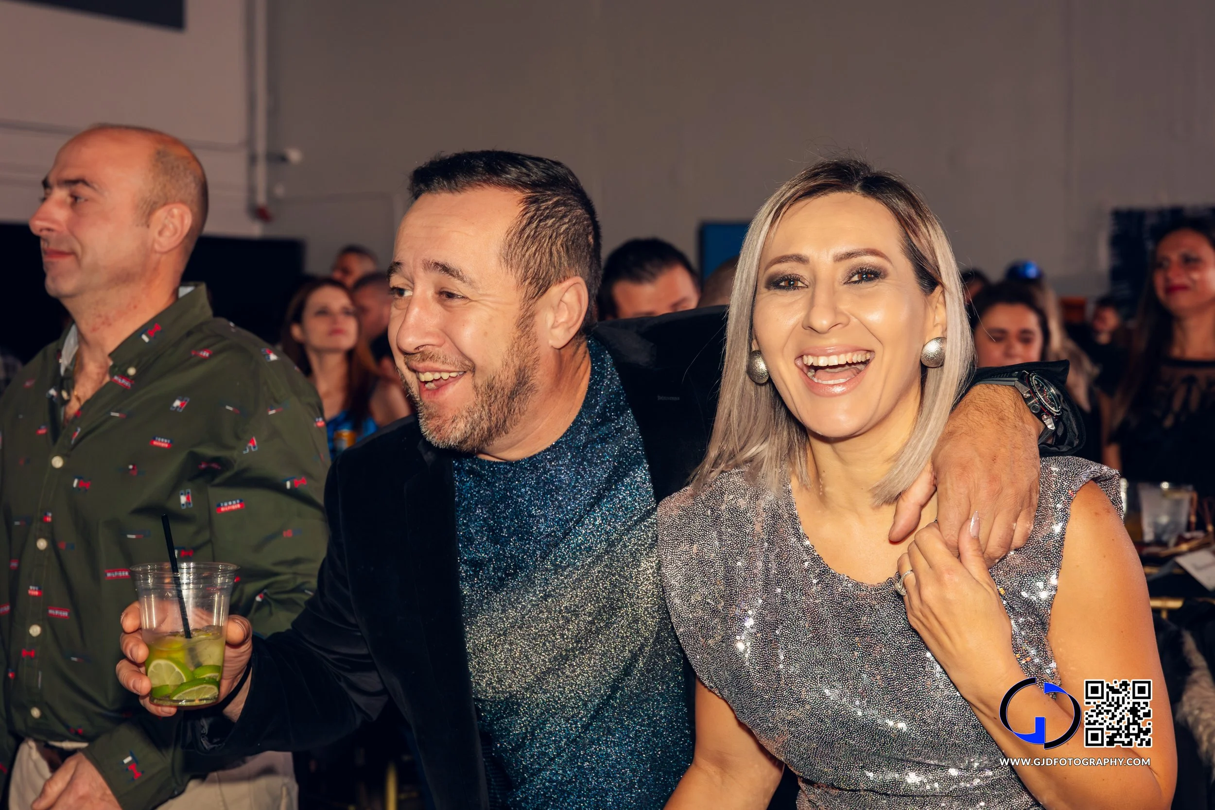 People enjoying a party, with a man holding a drink and a woman smiling and wearing a silver dress, in a crowded indoor setting.