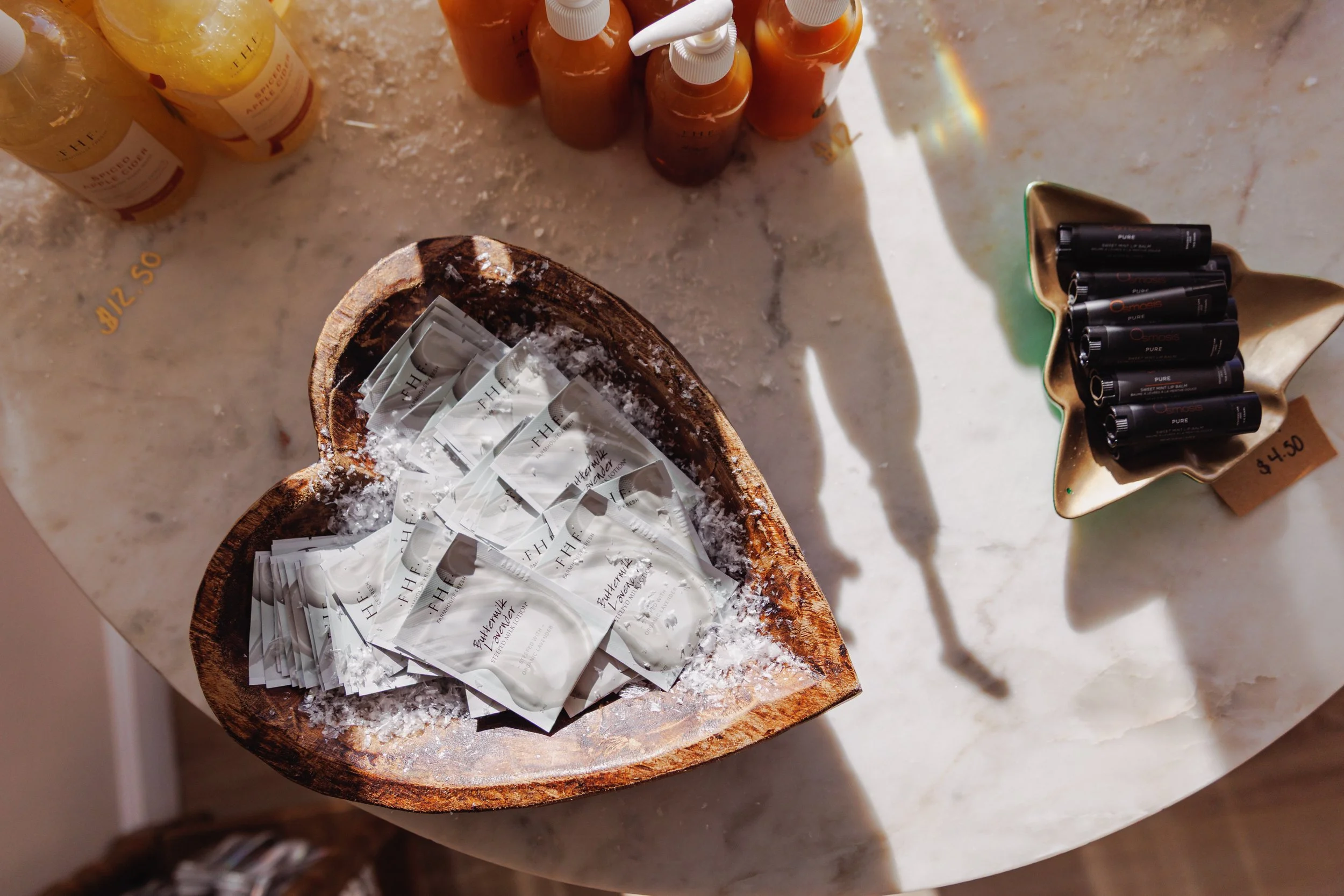 A marble table with randomly placed bottles of golden and orange liquids, a wooden dish holding several silver packaged items labeled 'Afterwork Defense,' and a leaf-shaped dish containing multiple black lipstick tubes, with a small price tag reading