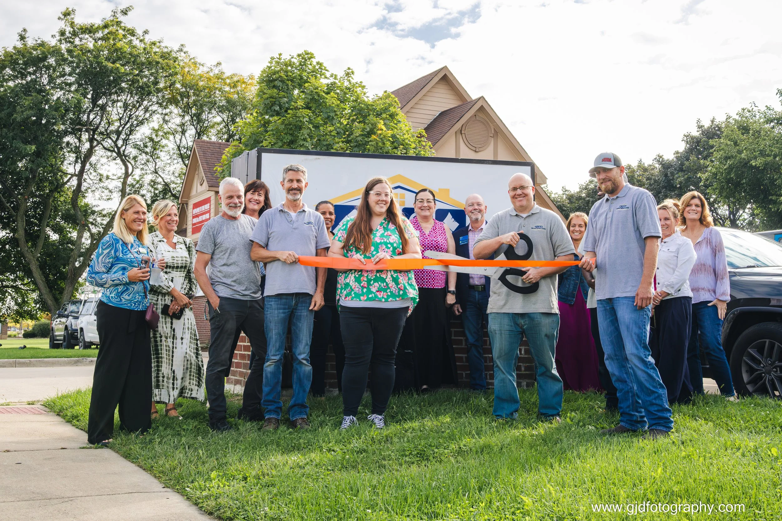 Group of people gathered outdoors for ribbon-cutting ceremony, standing in front of a house and a mobile home, with scissors and a ribbon in the foreground.