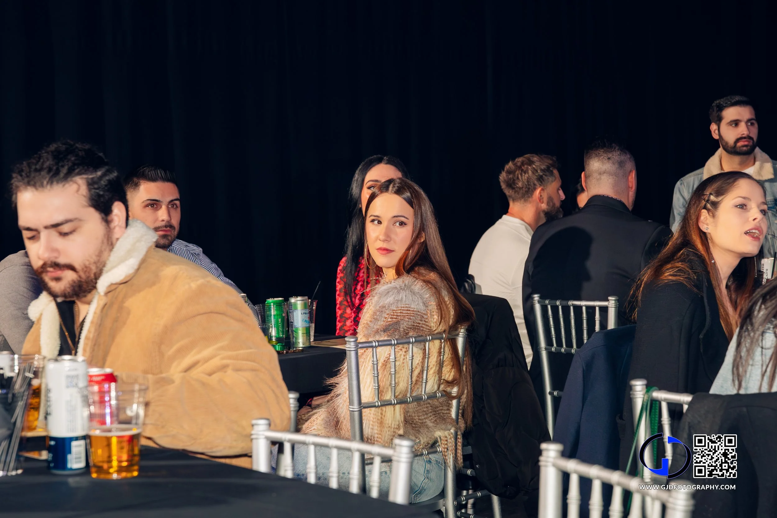 Group of people sitting at tables in a dimly lit event space, some looking towards the camera, some conversing, drinks and cans on the tables.