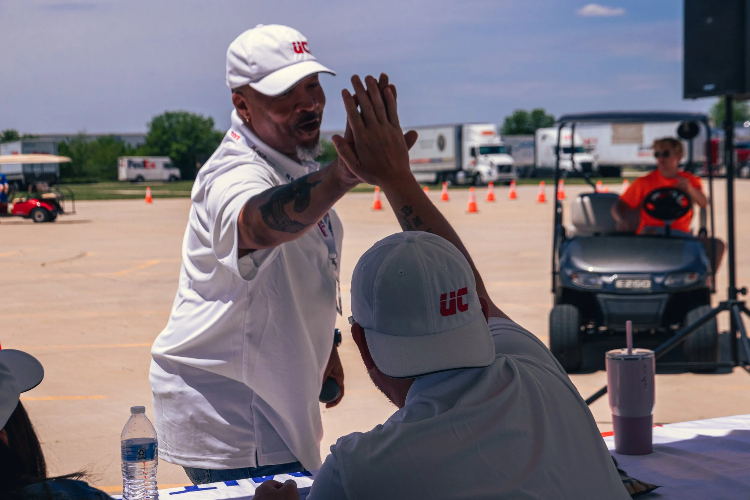 Two men, one with a cap and tattoos, giving a high-five. Both are wearing white shirts with 'UC' logos, outdoors on a sunny day with orange cones and trucks in the background.