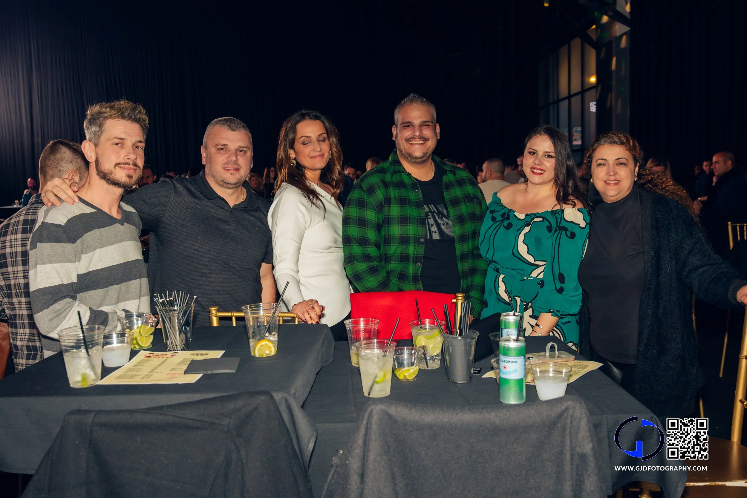 Group of six people standing behind a black table at a social event, smiling and posing for the camera.
