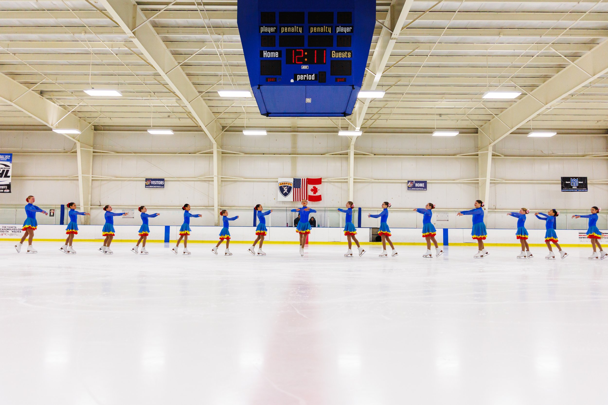 A group of synchronized figure skaters in matching blue costumes with yellow and red accents, skating in formation inside an indoor ice rink.
