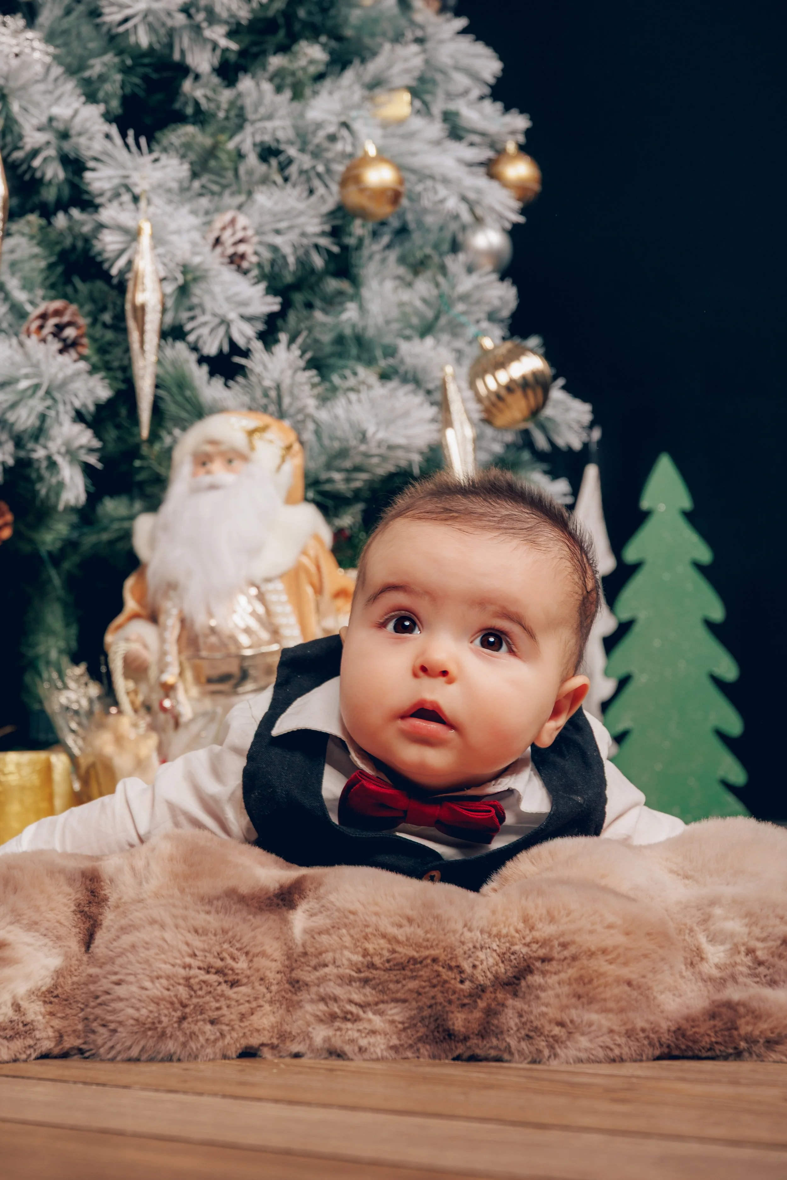 A baby in formal attire with a red bow tie, lying on a furry blanket in front of a decorated Christmas tree with ornaments and a Santa Claus figurine in the background.