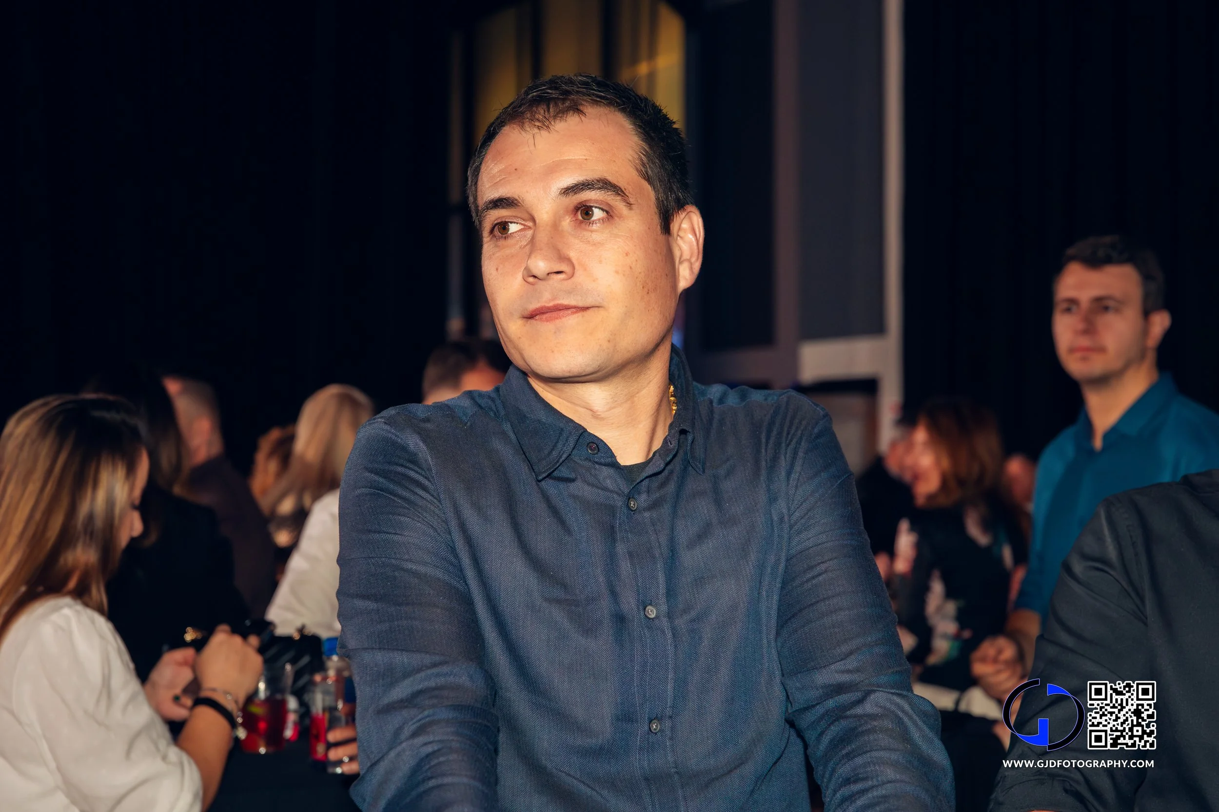 A man in a blue button-up shirt sitting with arms crossed at a crowded indoor event.