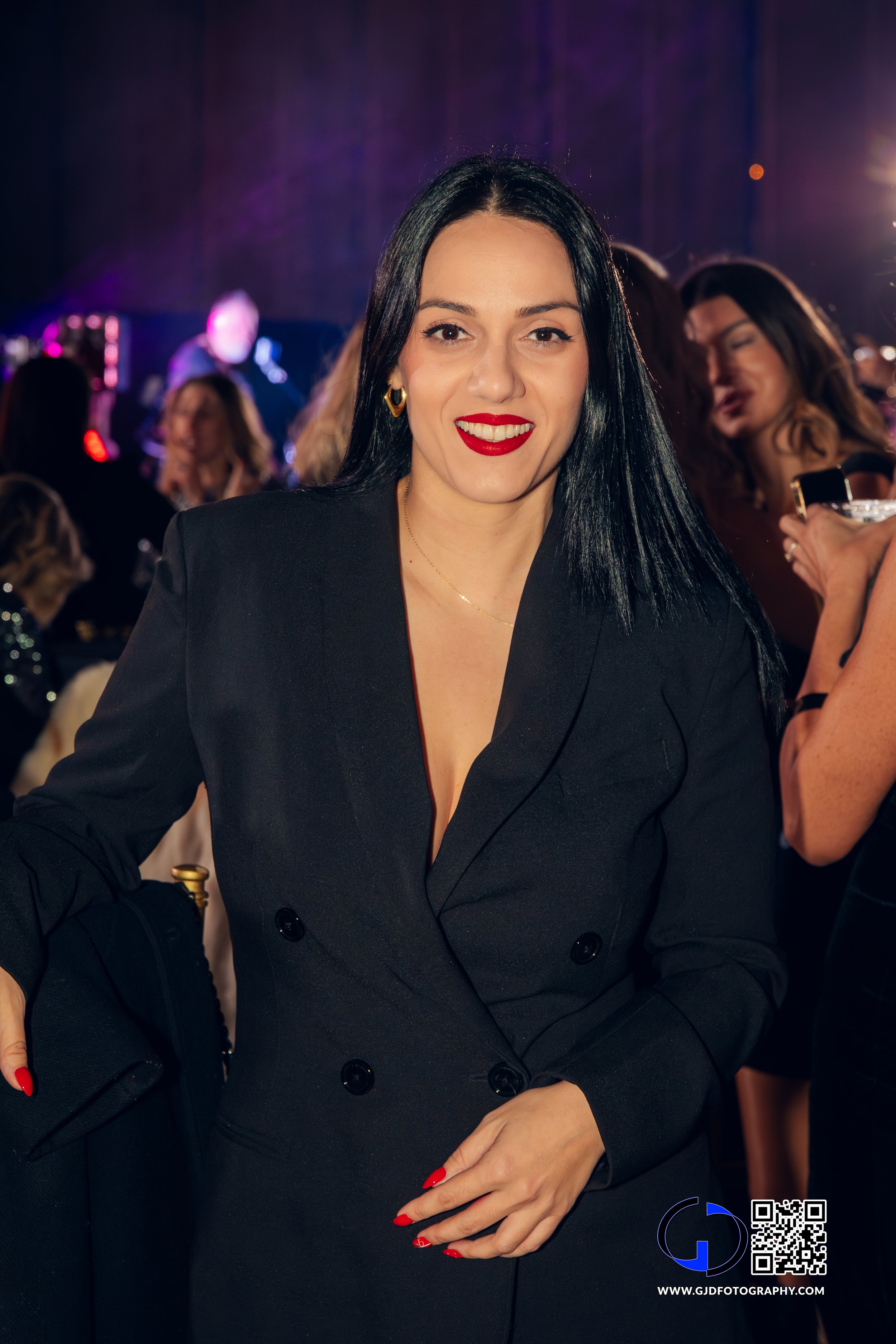 A woman with long black hair, wearing a black blazer with red lipstick, posing at a social event with other people in the background.