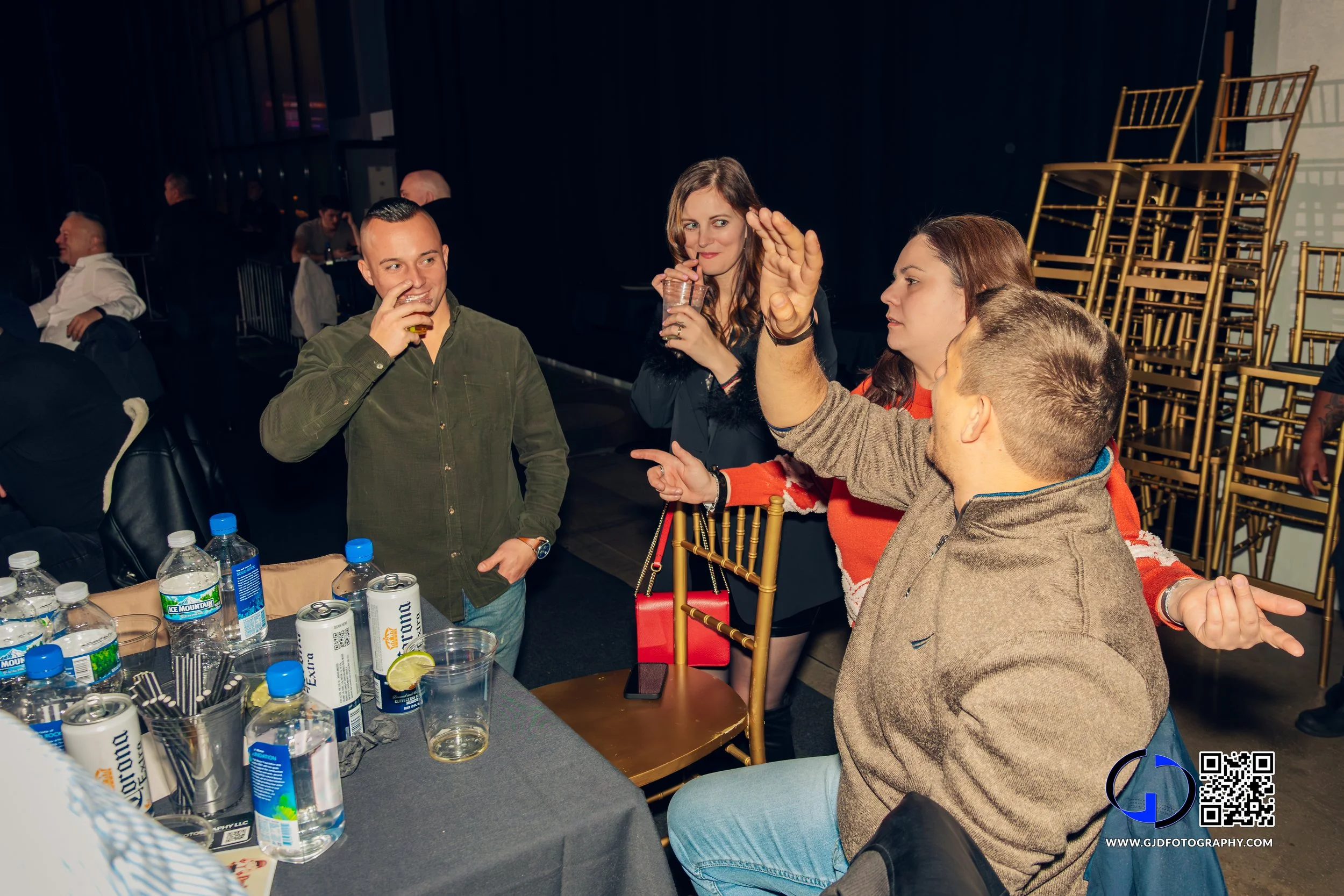 Group of four young adults socializing at a table with beverages in a dimly lit indoor setting, with stacked gold chairs in the background.