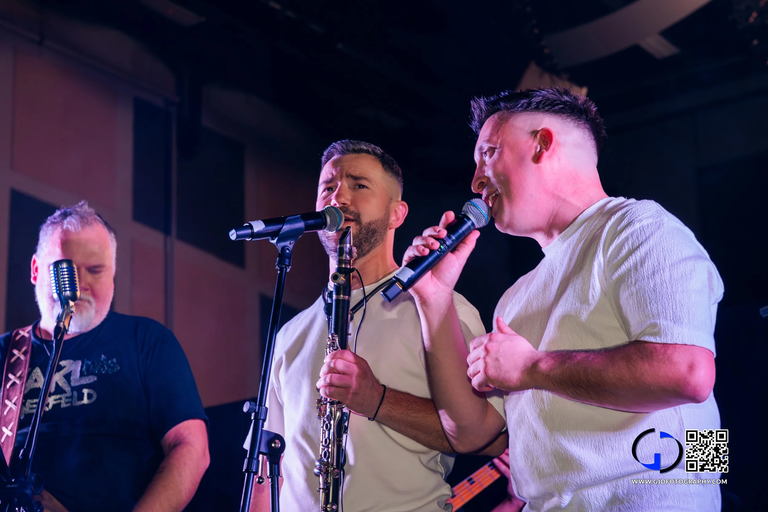 Three male musicians performing on stage with microphones, one playing a saxophone, under stage lighting.