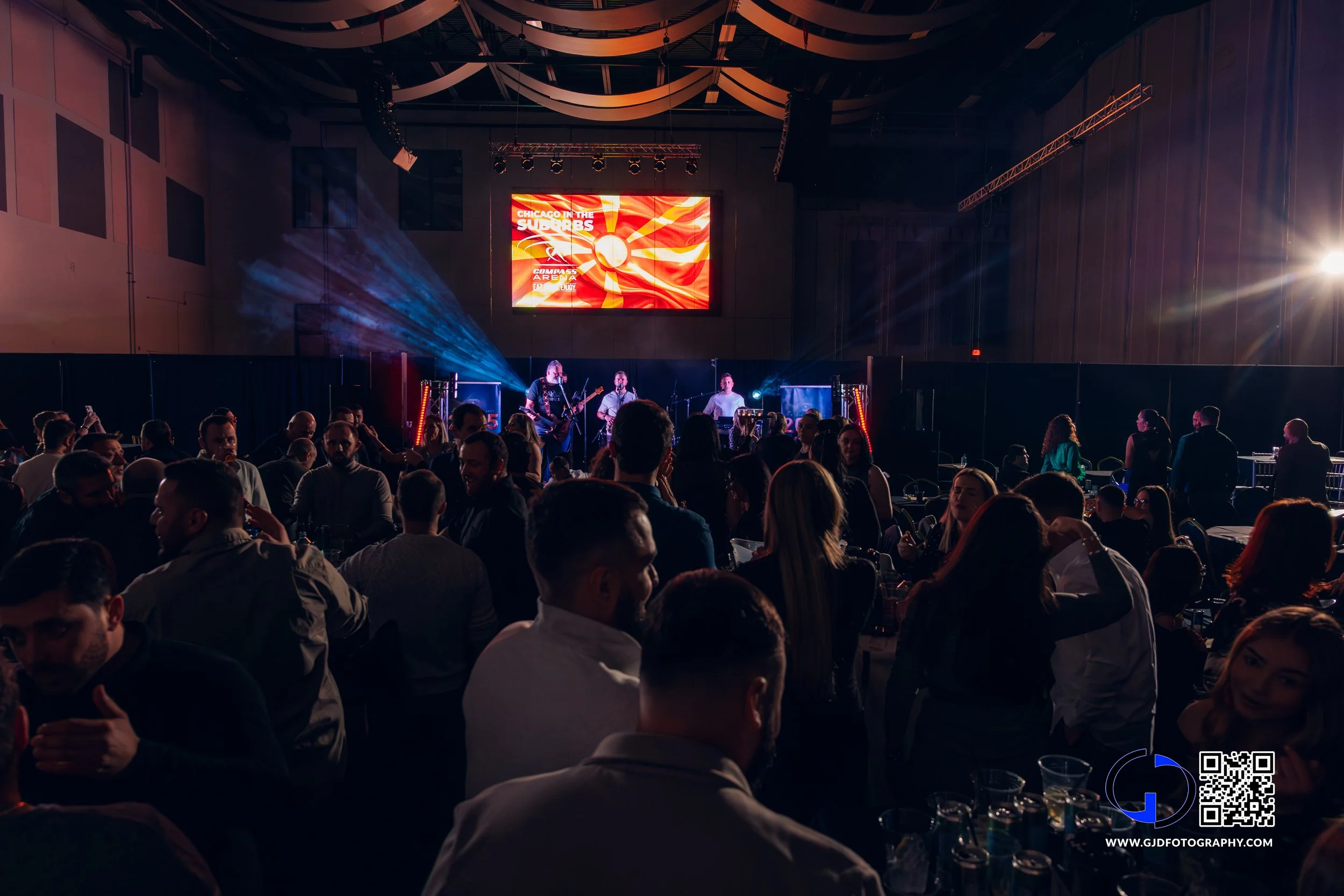 Crowd of people attending a live music concert indoors, with a band performing on stage under colorful lights and a large screen displaying red and yellow graphics in the background.