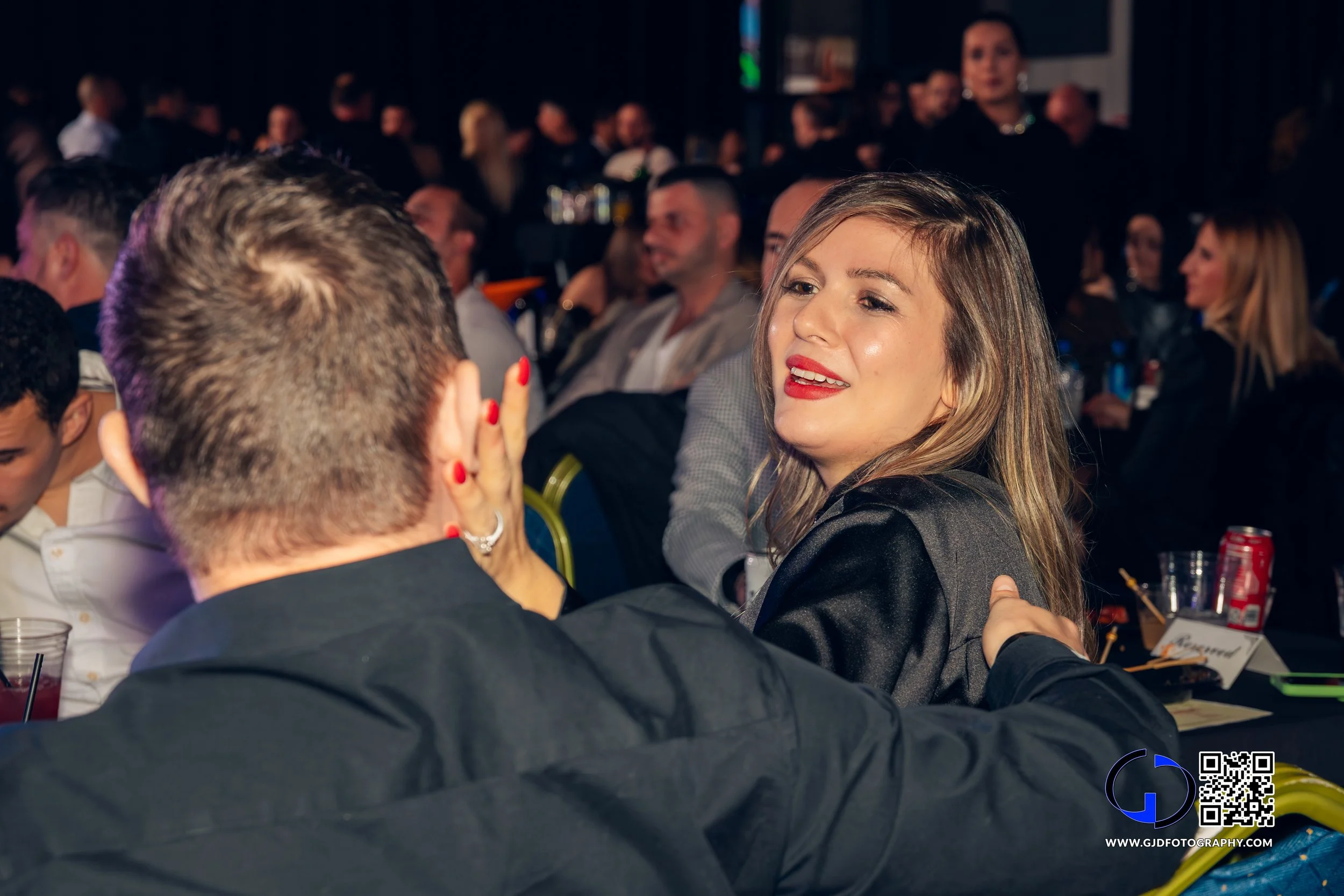 A woman with blonde hair and red lipstick is talking with a man at a crowded event, with other people seated at tables in the background.