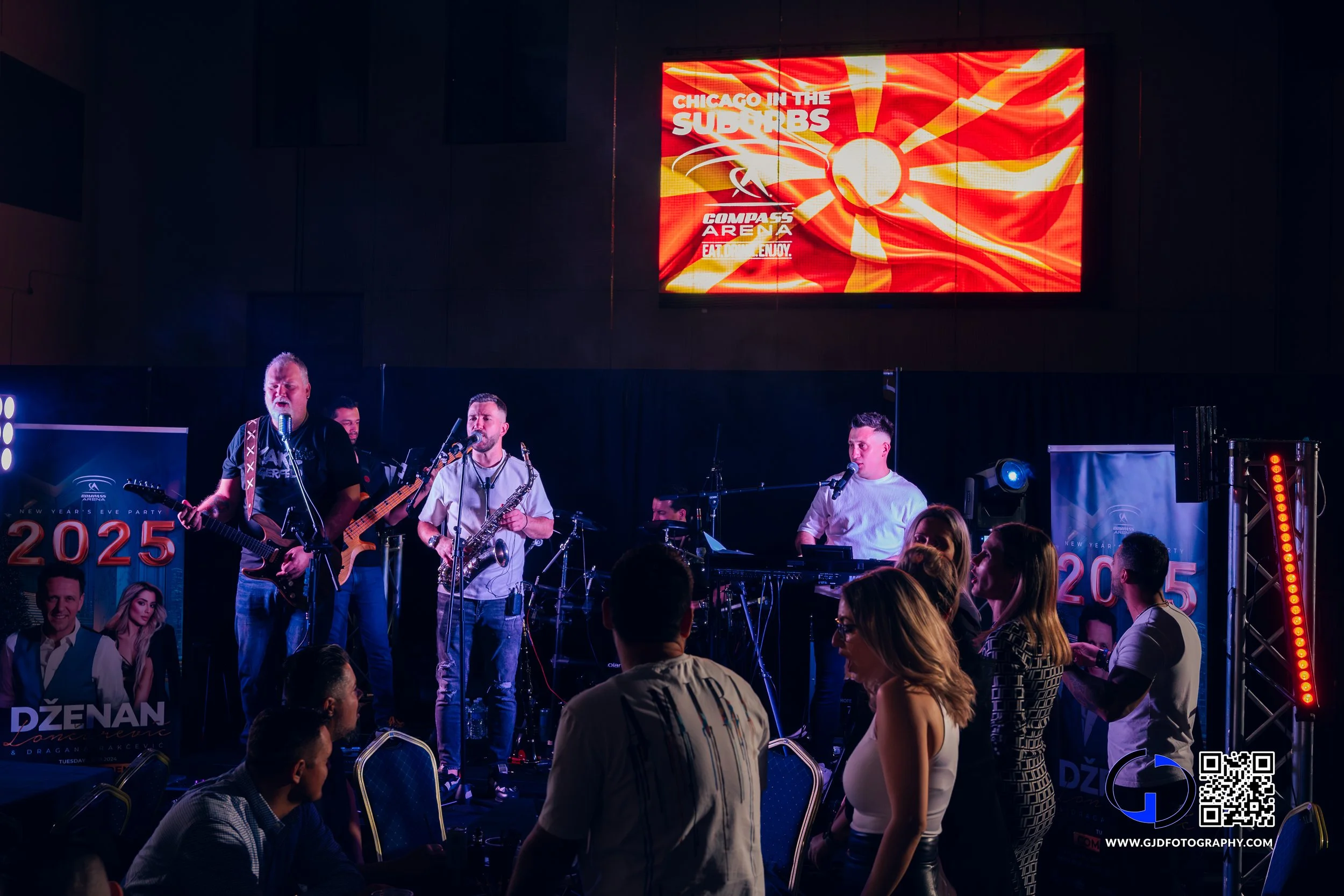 A band performs live on stage at a New Year's Eve party, with a 2025 banner, attendees watching, and a large screen displaying a red and yellow graphic.