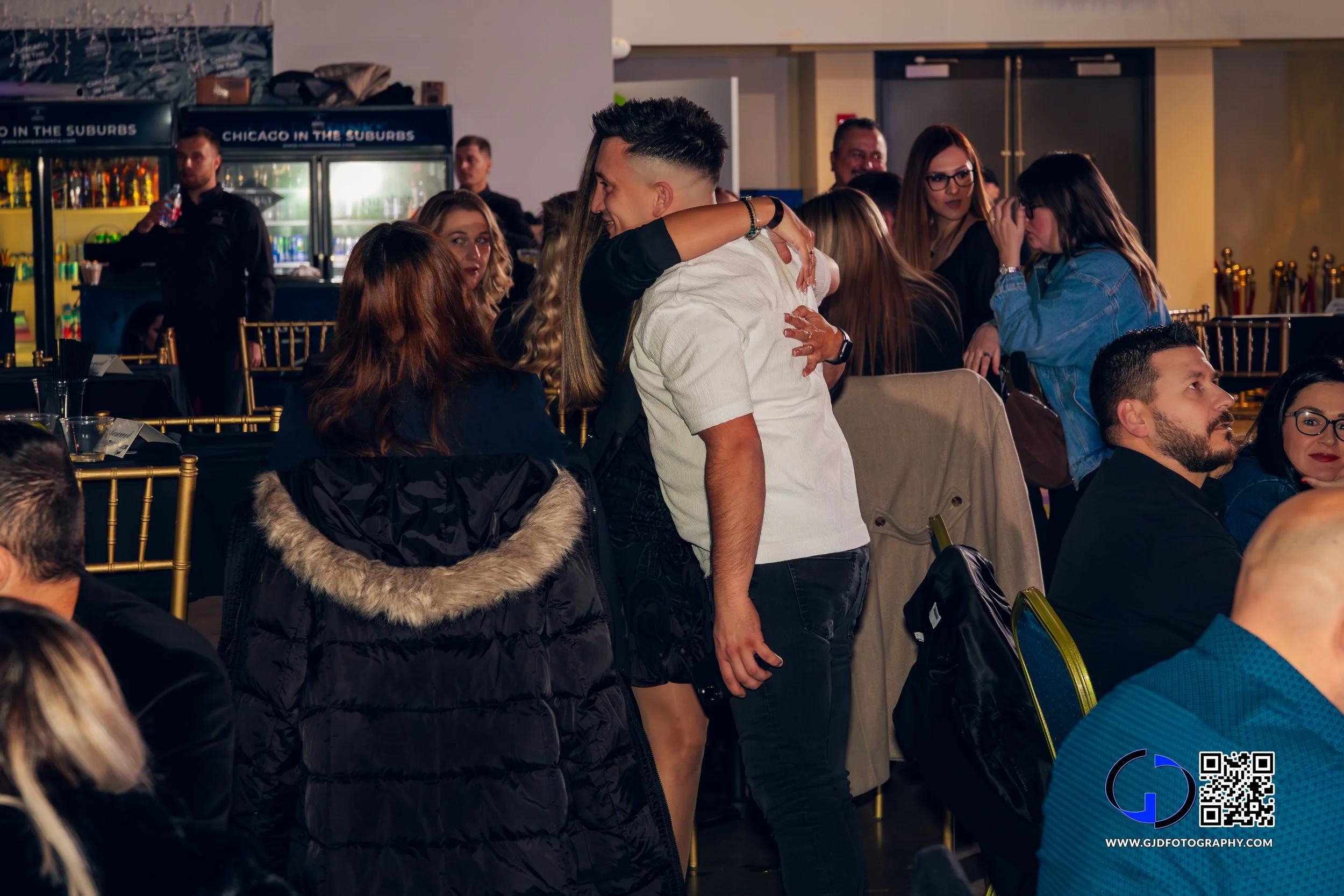 A group of people at a social gathering, some are sitting at tables while others are standing and hugging in an indoor venue with gold chairs, a DJ booth, and a colorful bar in the background.