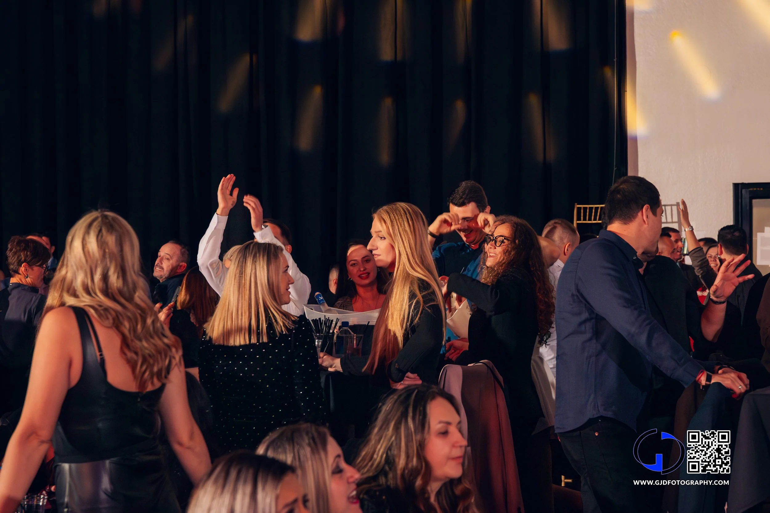 Crowd of people dancing and talking at a lively indoor event or party, with a black curtain background.