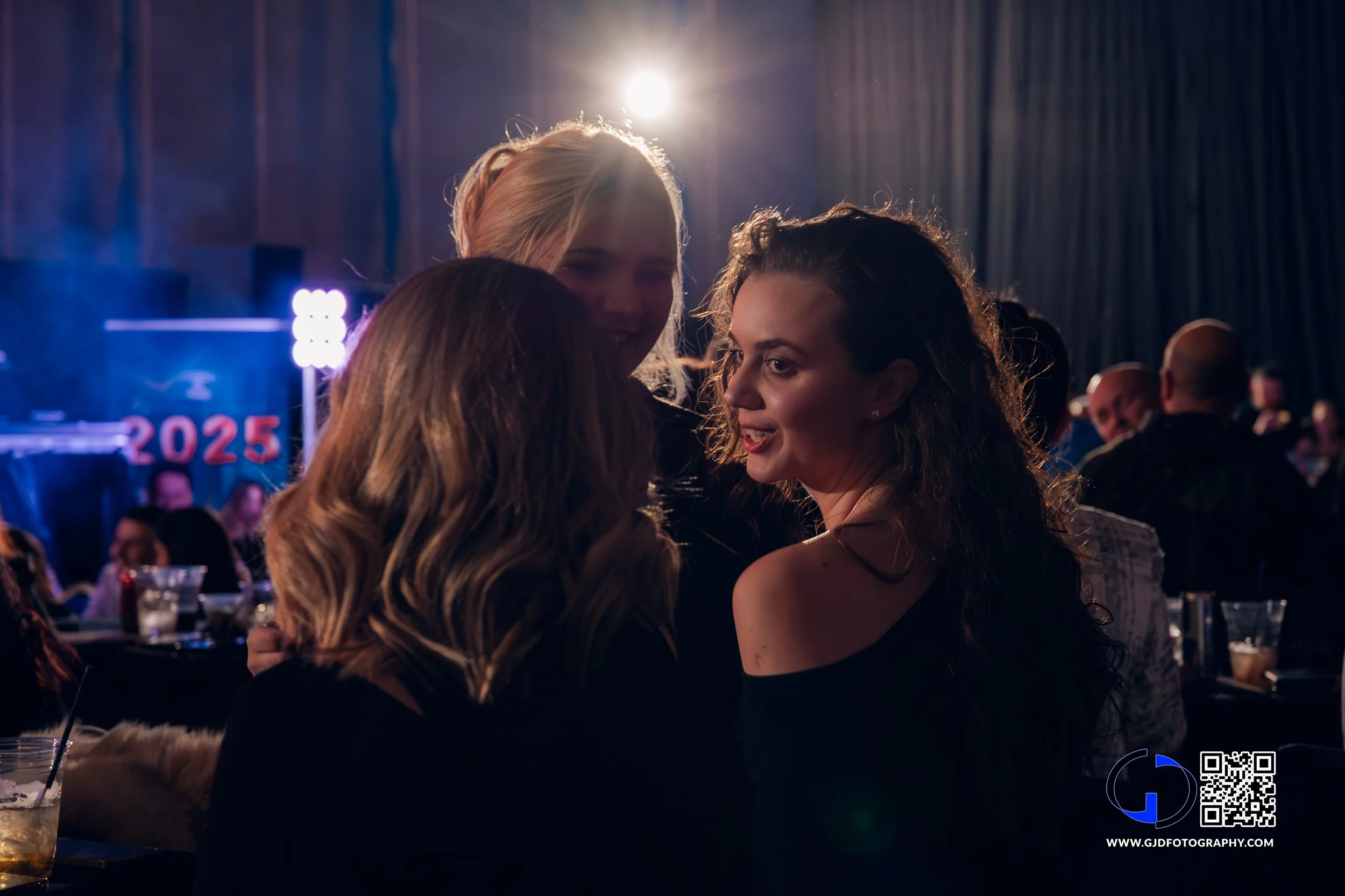 Three women smiling and talking at a celebration event with '2025' in the background and a DJ setup, illuminated by stage lights.