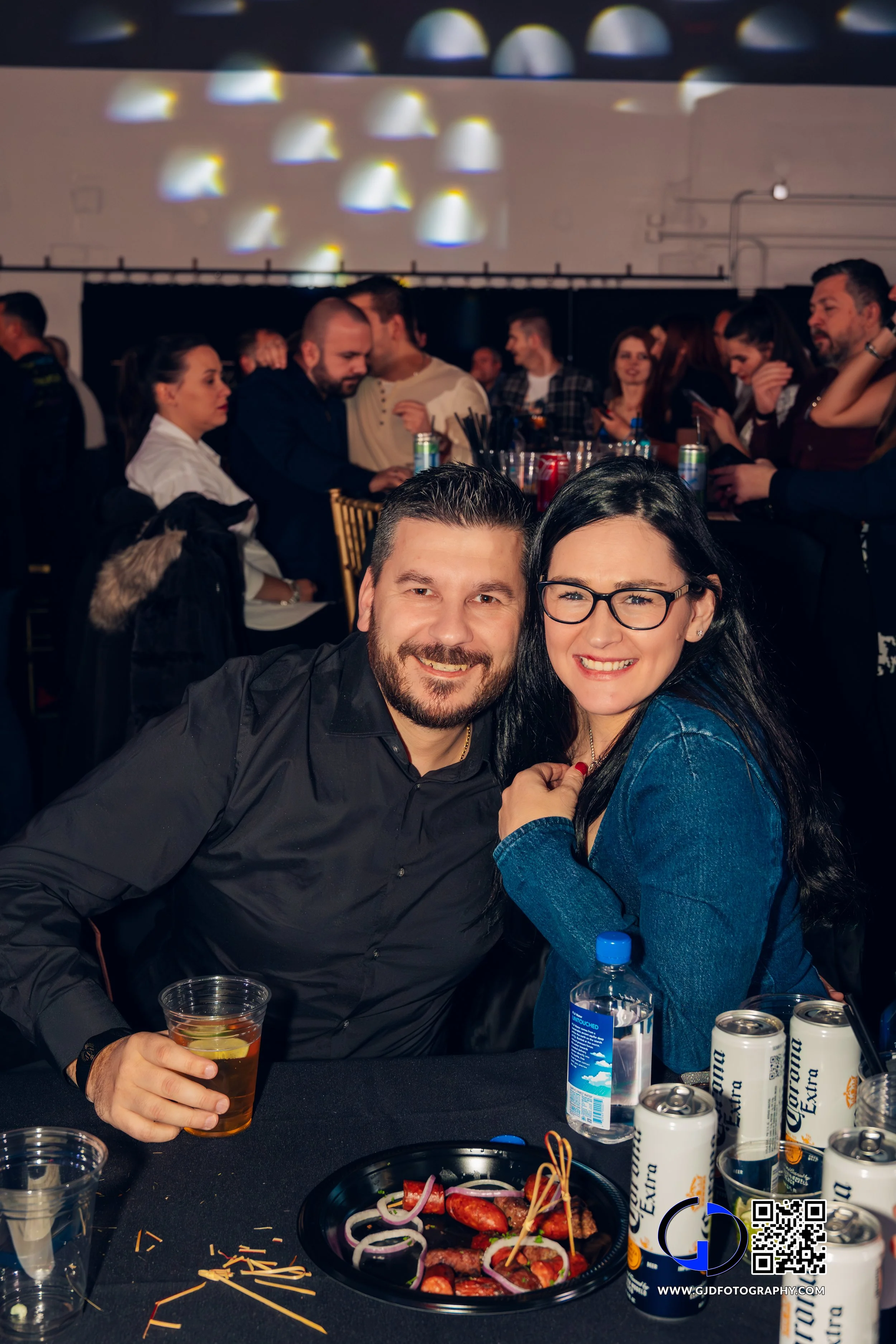A smiling couple seated at a table with drinks and food in a lively, crowded indoor event or party.