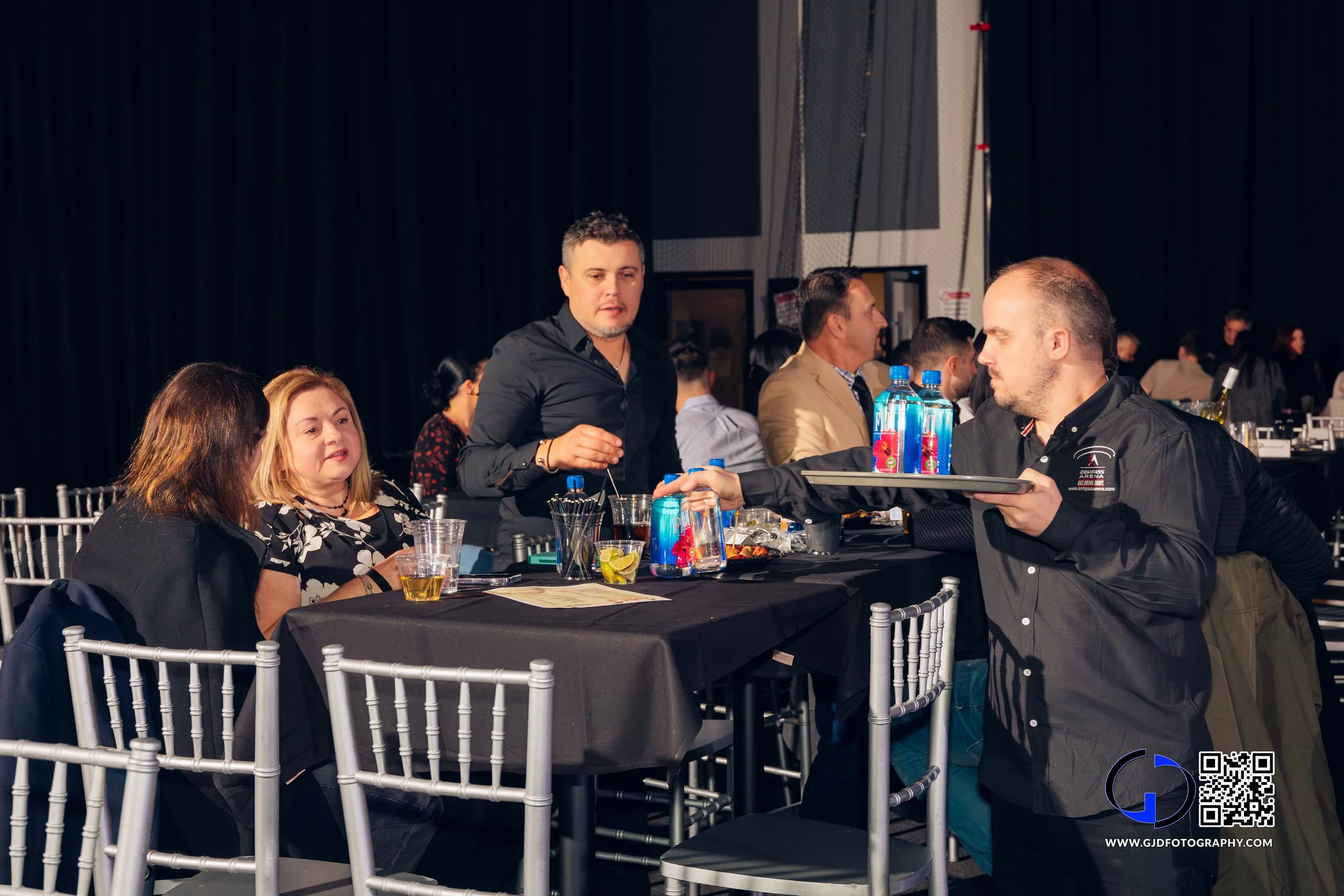People sitting at a formal event, one person serving drinks to two women seated at a table with black tablecloths and silver chairs