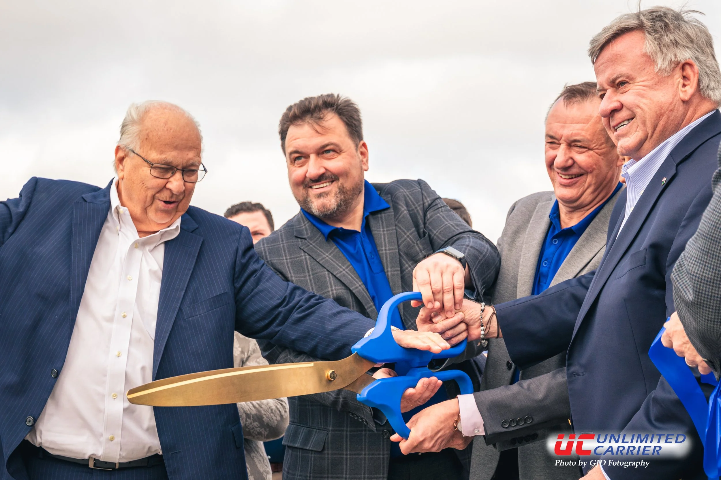 Group of men in suits and blazers holding large ceremonial scissors, smiling, at an outdoor event during daytime, with cloudy sky in background.