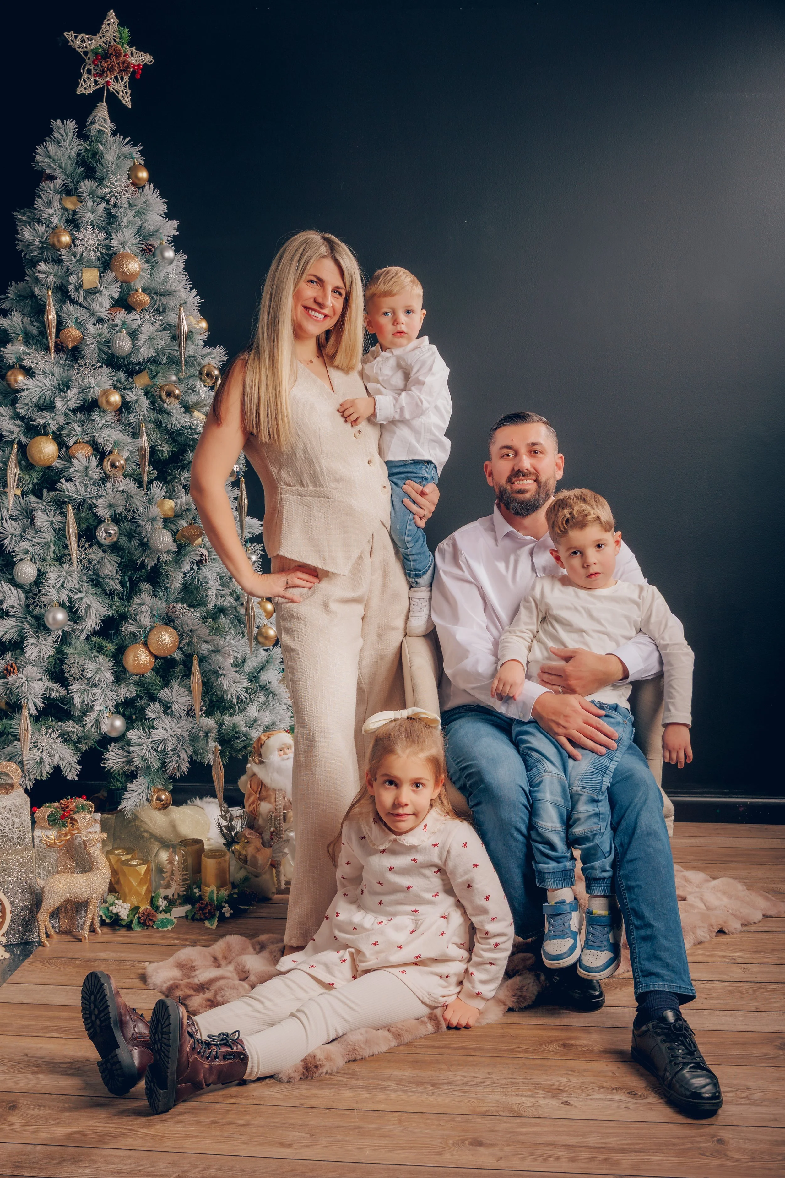 A family of five posing indoors next to a decorated Christmas tree. The mother stands with two children, a boy and girl, while the father sits with another boy on his lap. The scene is festive with Christmas decorations and gifts visible.