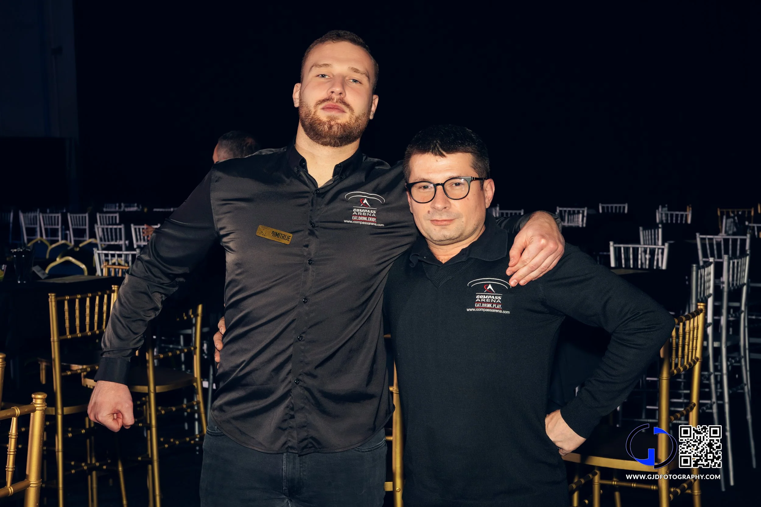 Two men standing together at an event, wearing black shirts with logos, surrounded by empty gold chairs, one with a beard and the other with glasses.