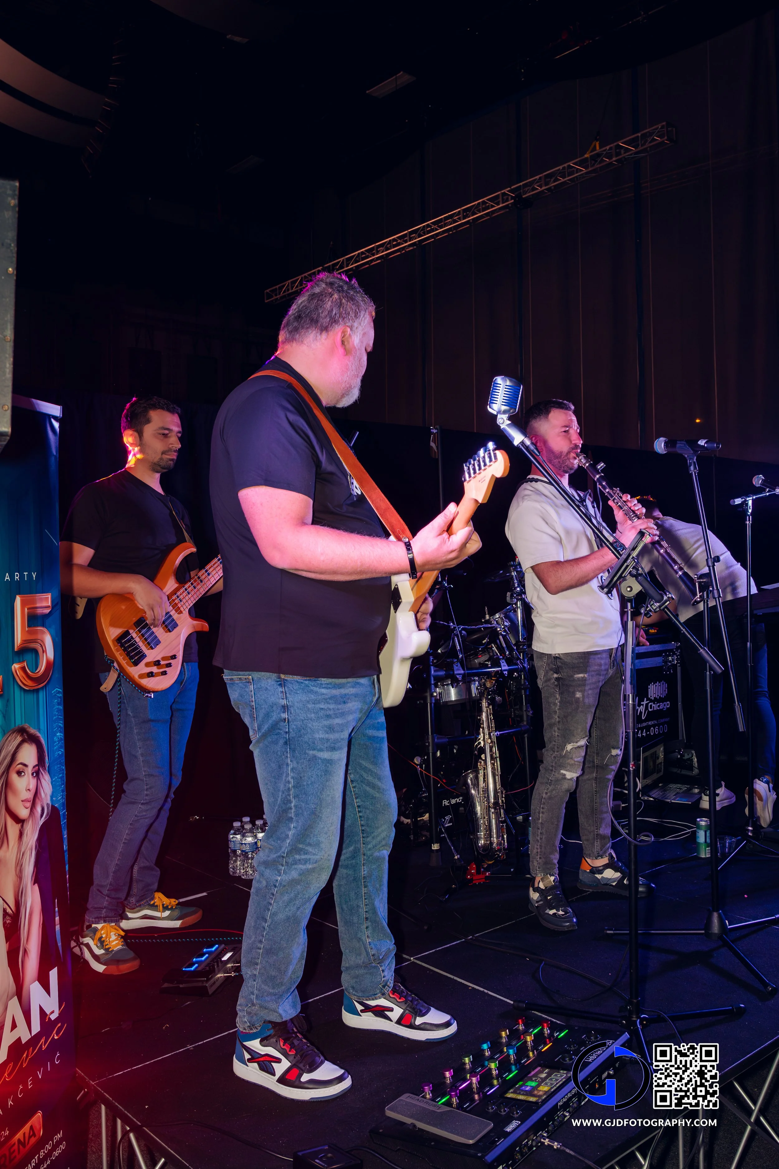 A band of four musicians performing on stage, playing guitars, bass, and wind instruments under stage lighting.