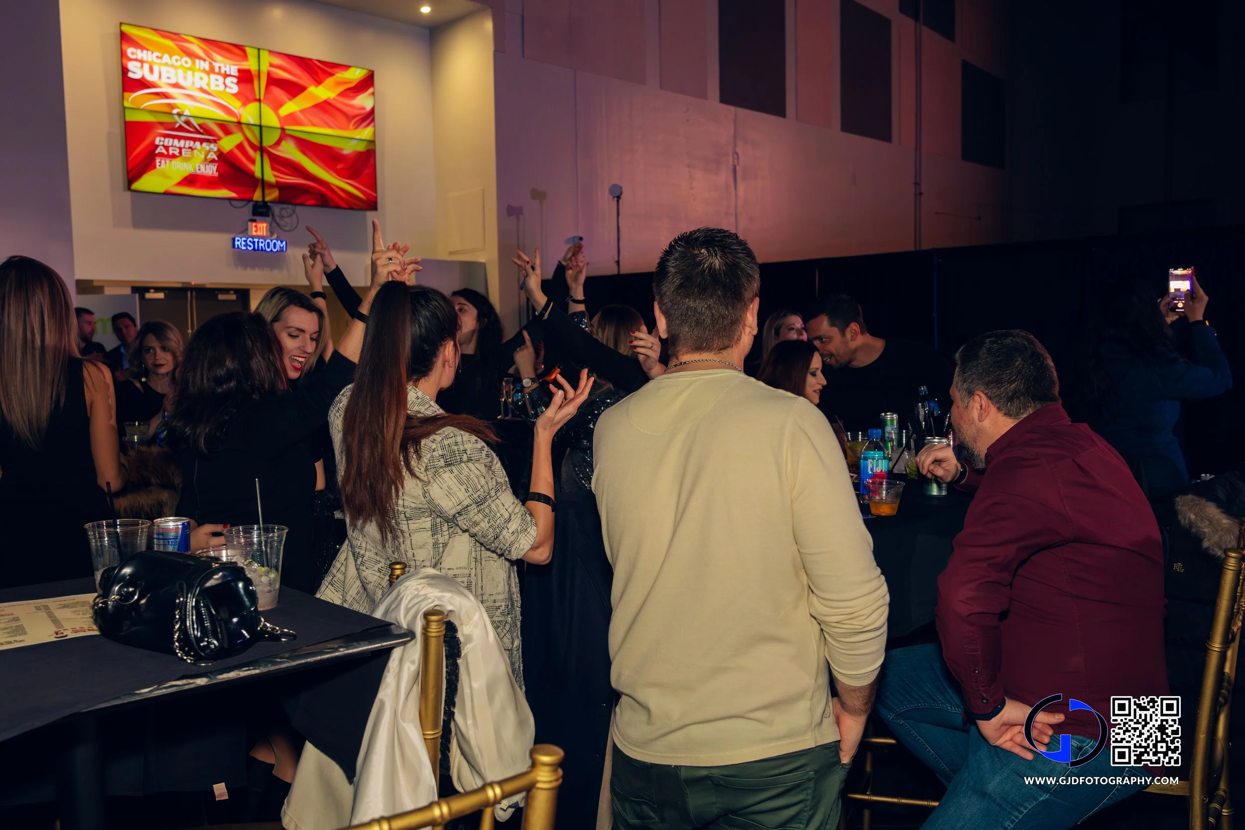 People enjoying a party in a dimly lit room with a large screen displaying "Chicago in the Suburbs" in the background, some dancing and others taking photos.