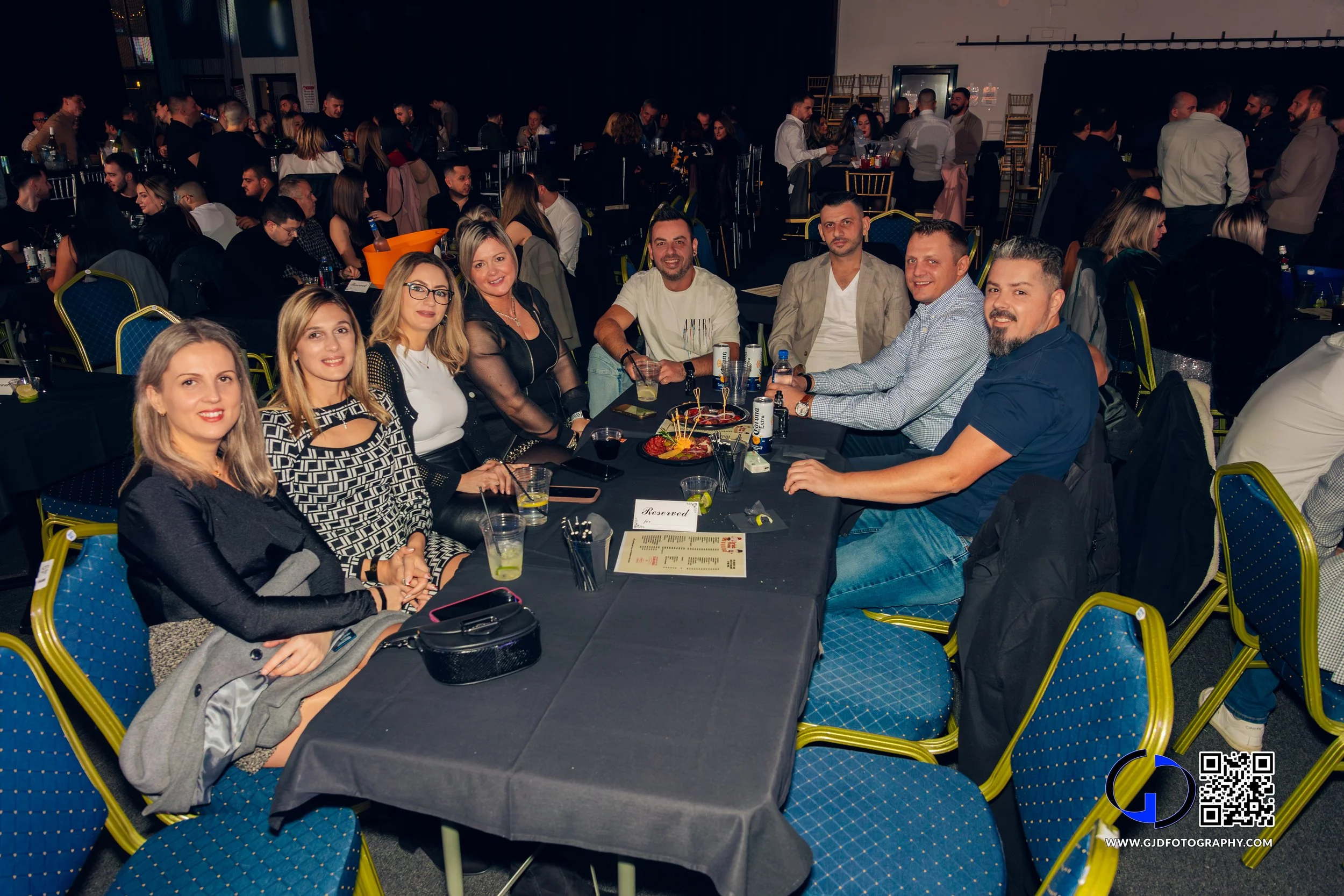 A group of nine people sitting at a black-covered table during a social event or party, with drinks and food, in a large indoor venue filled with other guests and tables.