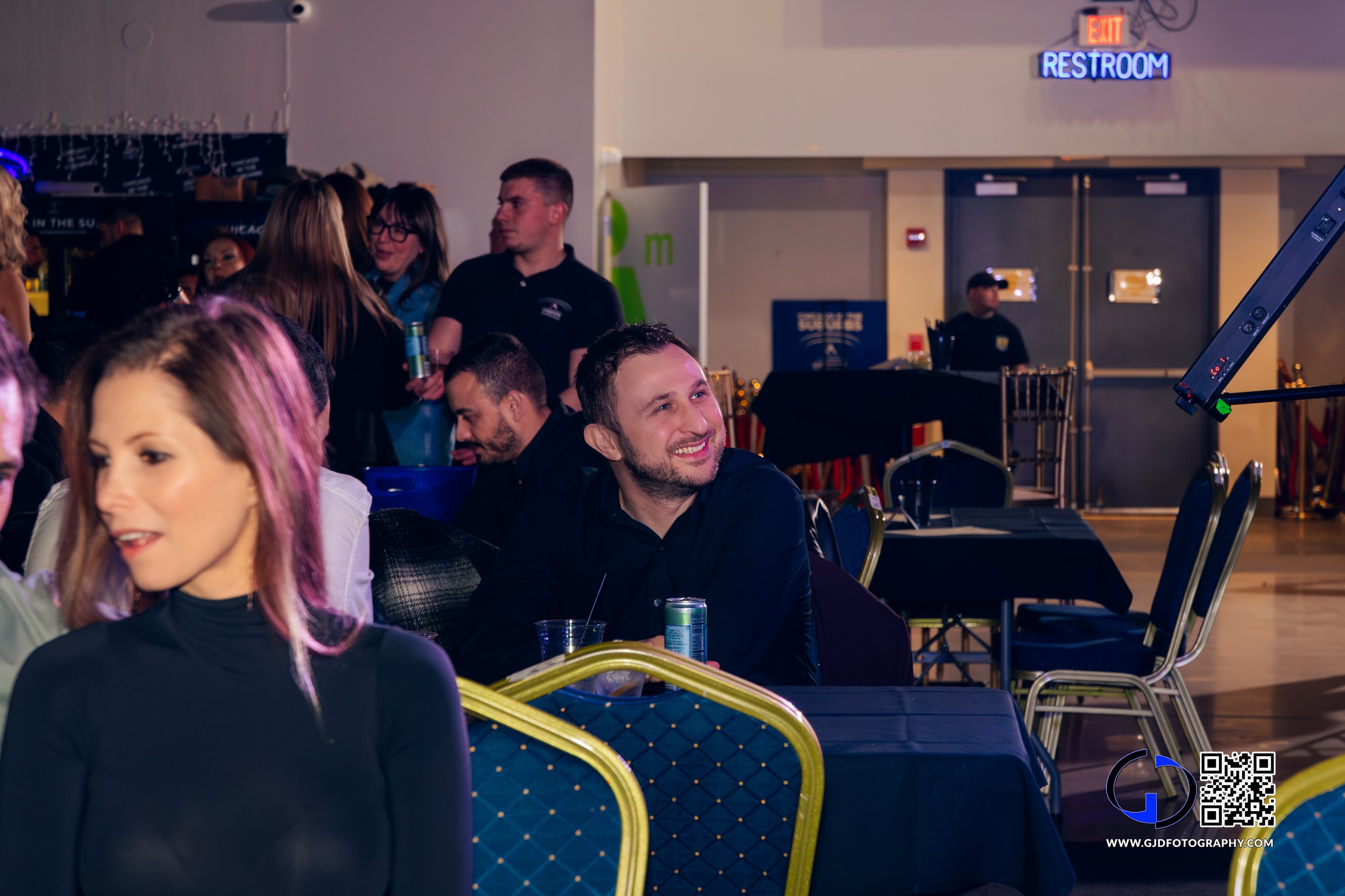 People socializing at an indoor event with tables and chairs, a man smiling sitting at a table with a drink and a can, and a neon exit sign and restroom sign in the background.