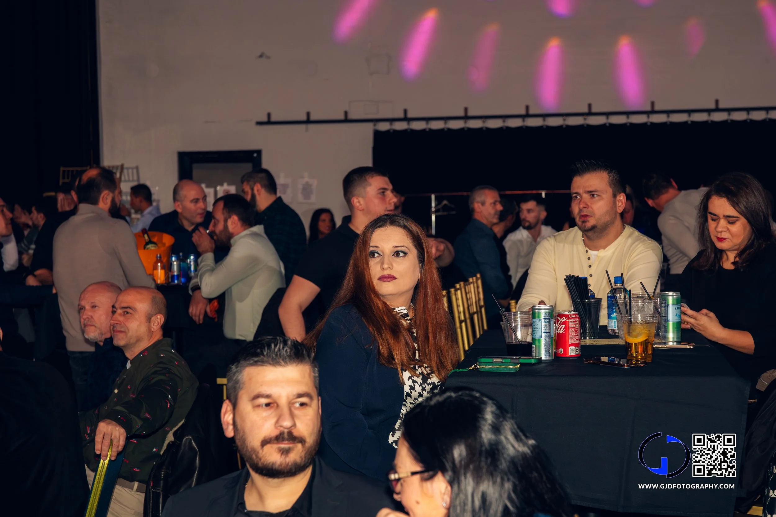 People attending a social event or party, seated at tables with drinks, in a dimly lit room with purple and pink lighting effects.