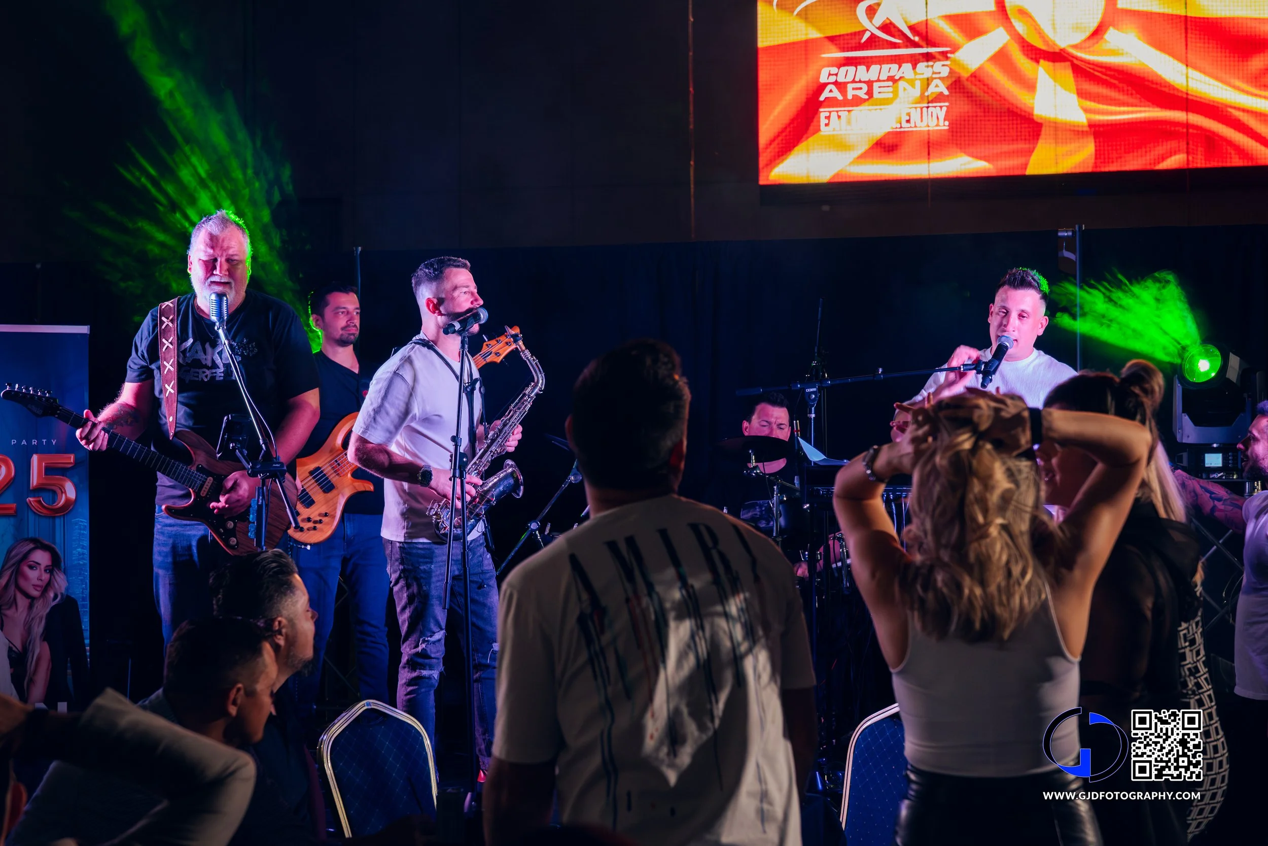 Live band performing on stage at Compass Arena with audience dancing and enjoying the show, colorful stage lighting, large screen displaying concert info.
