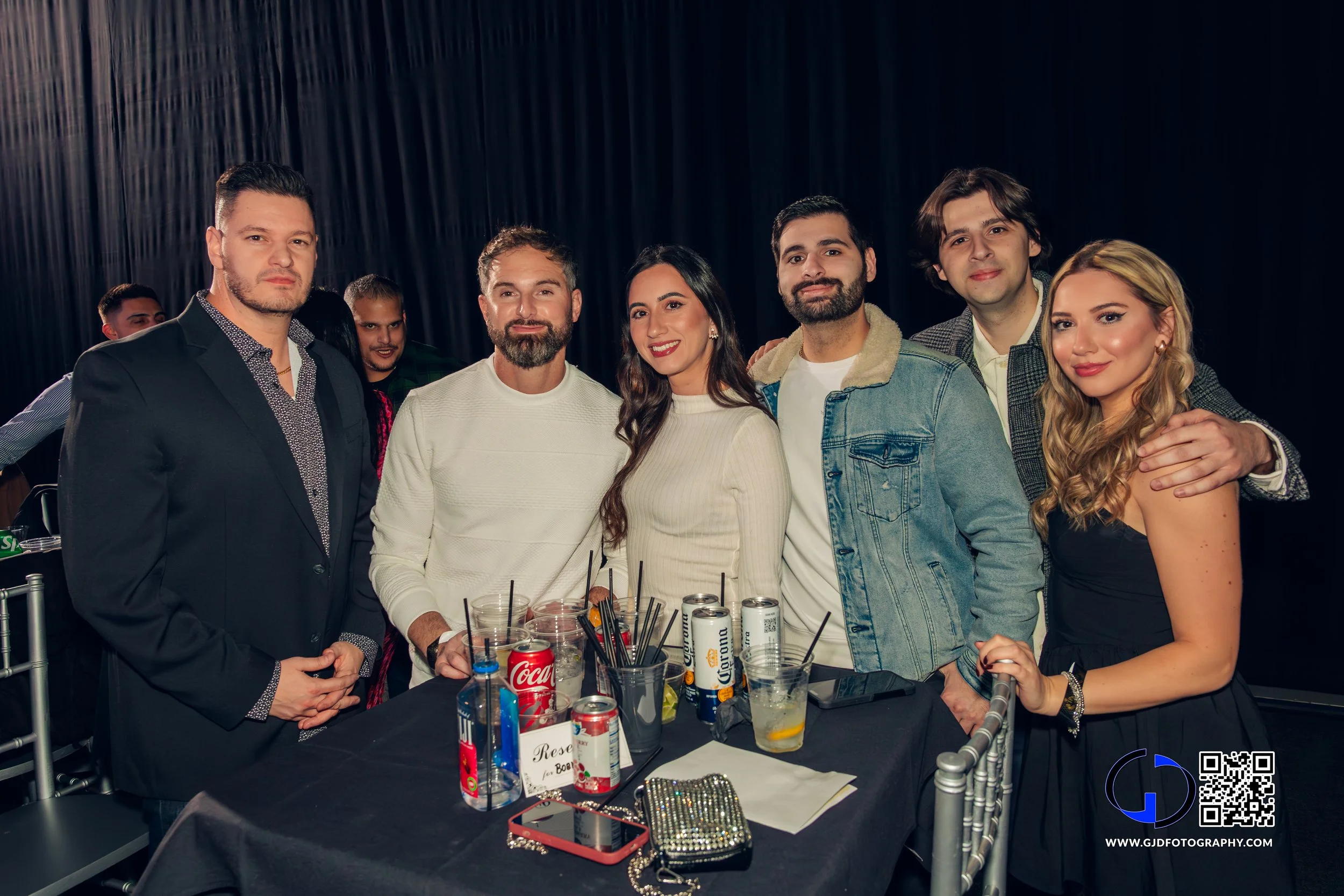 Group of seven young adults gathered around a table at a party or event, smiling at the camera, with drinks and party supplies on the table, and a dark curtain backdrop.