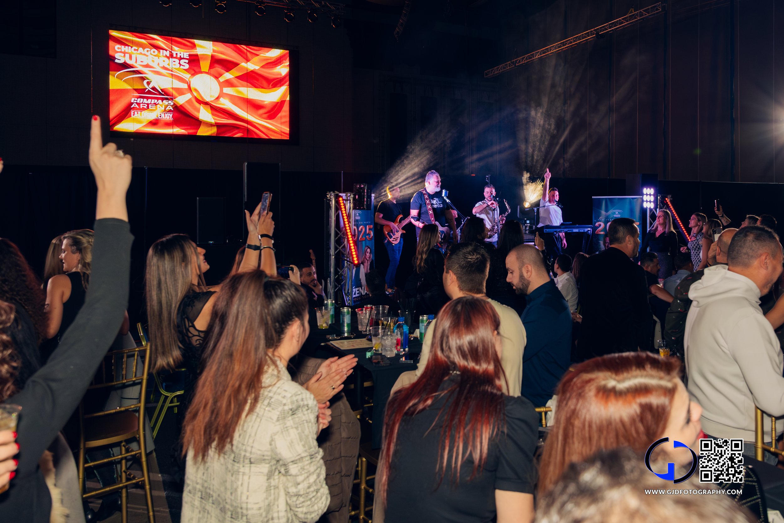 Audience watching a live band performance on stage with large screen above displaying 'Chicago in the Suburbs' and event details, in an indoor venue.