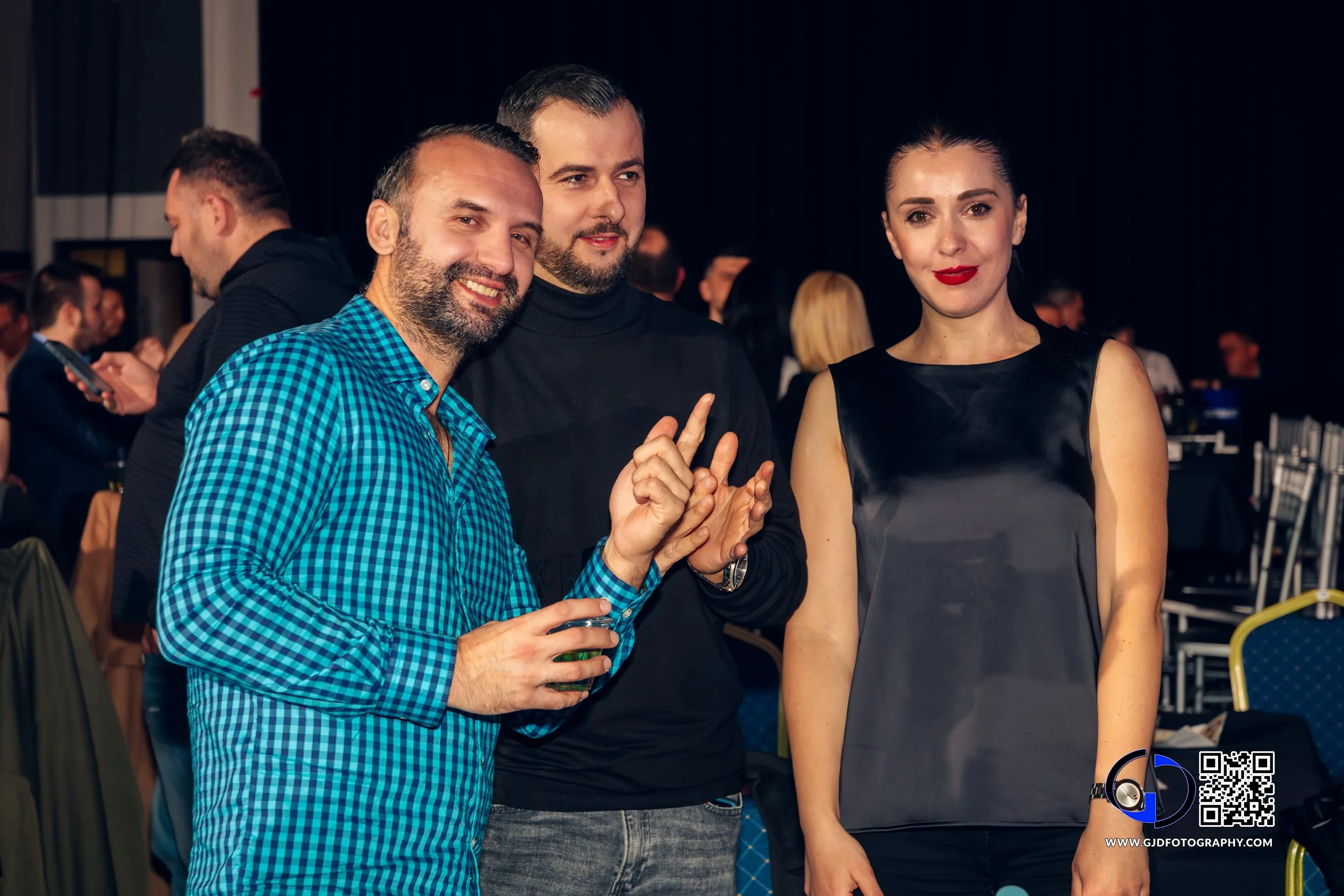 Three people posing together at a social event, smiling, holding drinks, with a group of people in the background.