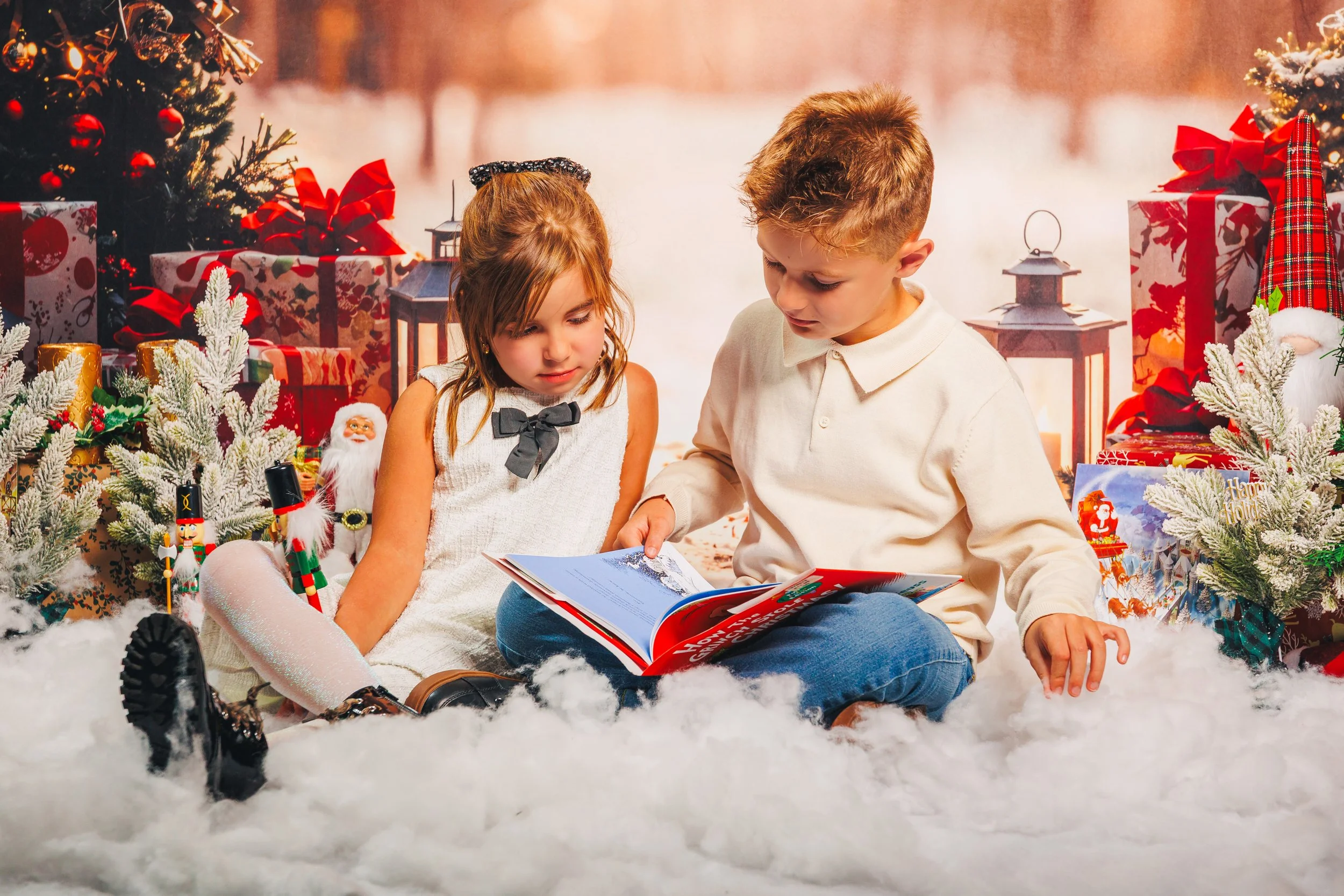 Two children, a girl and a boy, sitting on a fluffy white surface surrounded by Christmas decorations, including wrapped presents, Christmas trees, and ornaments, reading a book together with a festive background.