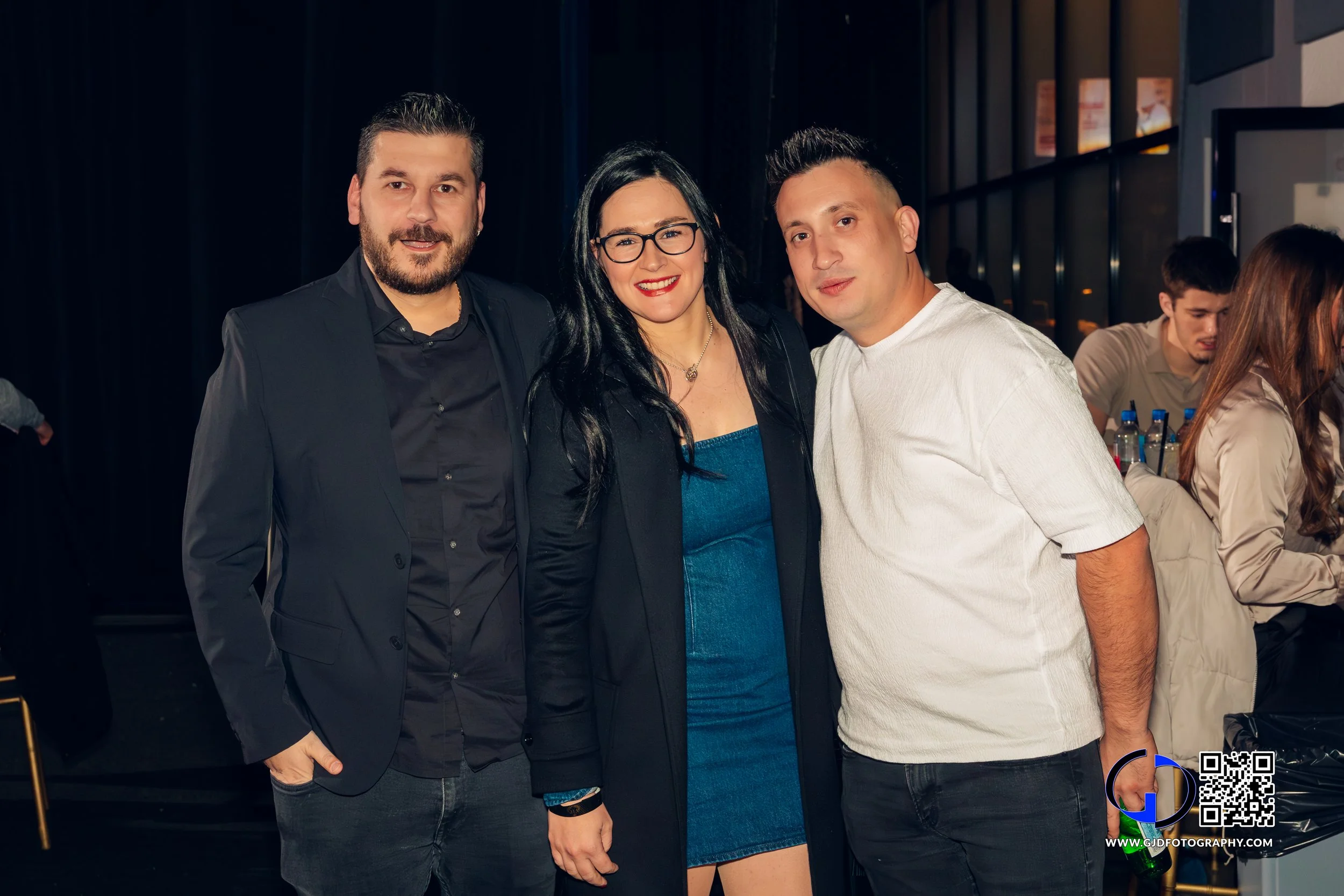 Three people posing together indoors, two men and one woman, smiling and dressed in semi-formal attire, with a bar and other people in the background.