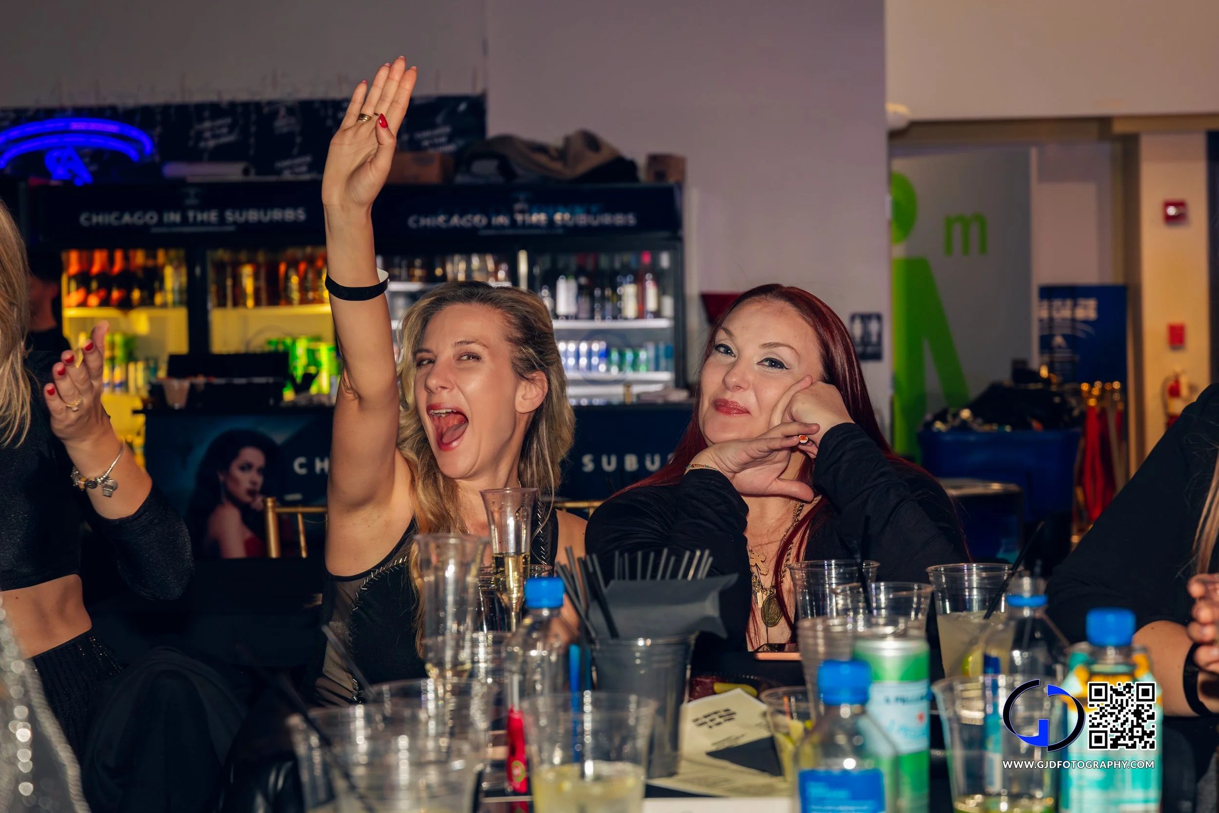 Two women sitting at a table in a bar or restaurant, one raising her hand and smiling, the other with a relaxed expression. The table has drinks and cups, with a bar in the background.