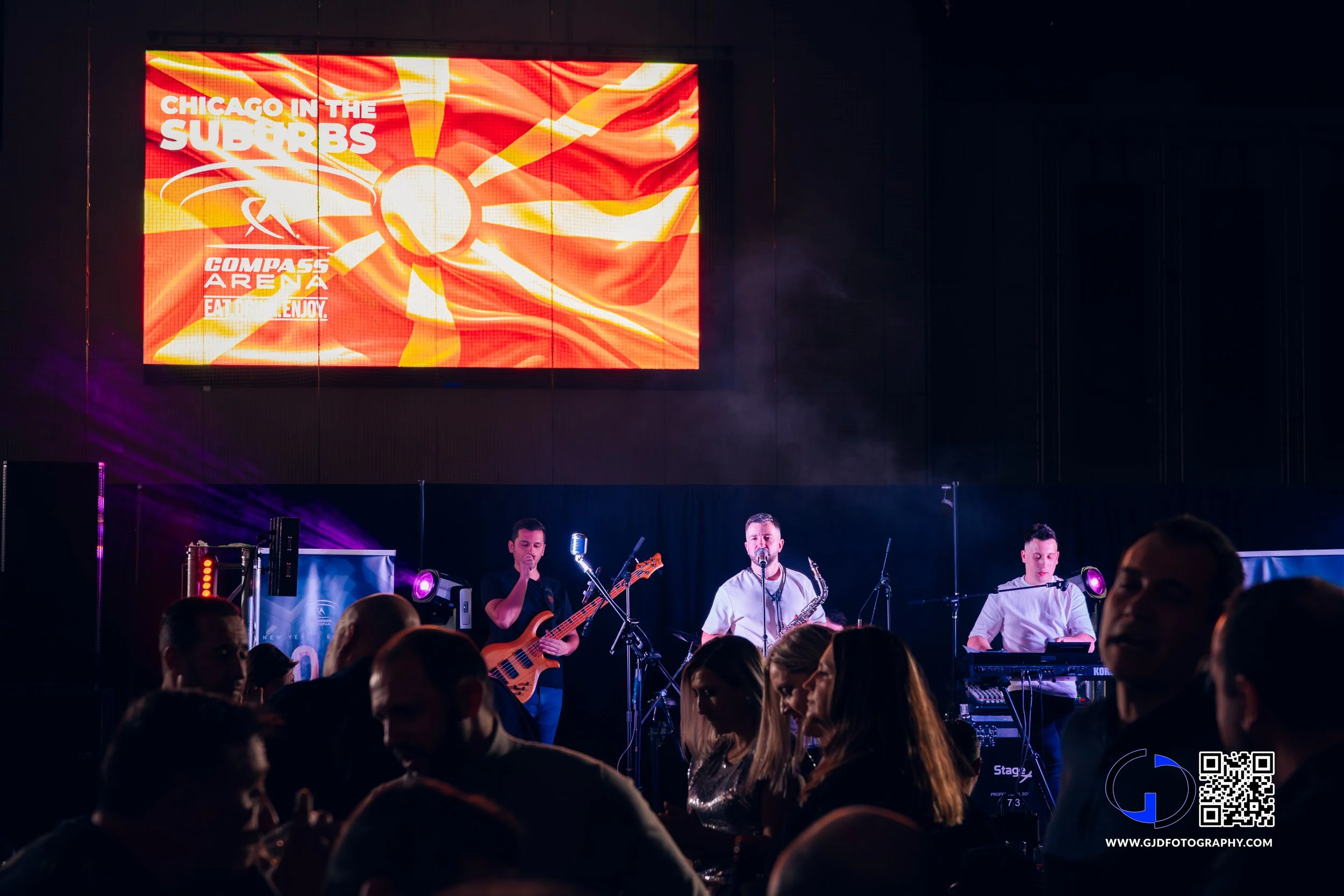 A live music performance on stage with three band members and an audience in front, with a large screen displaying an image of the Chicago flag and text that reads "Chicago in the Suburbs" and "Compass Arena."