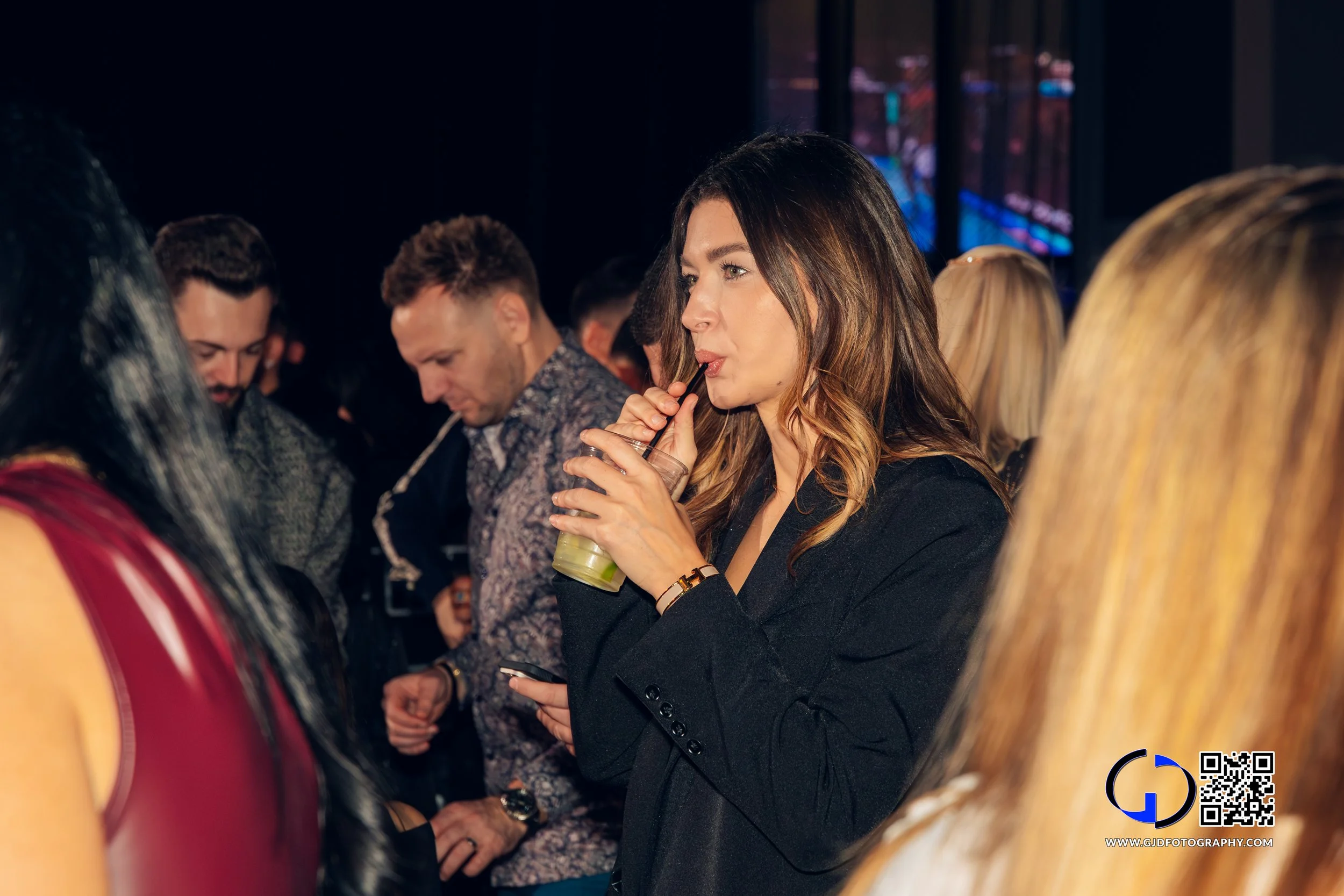 A woman drinking a beverage with a straw at a crowded indoor event.