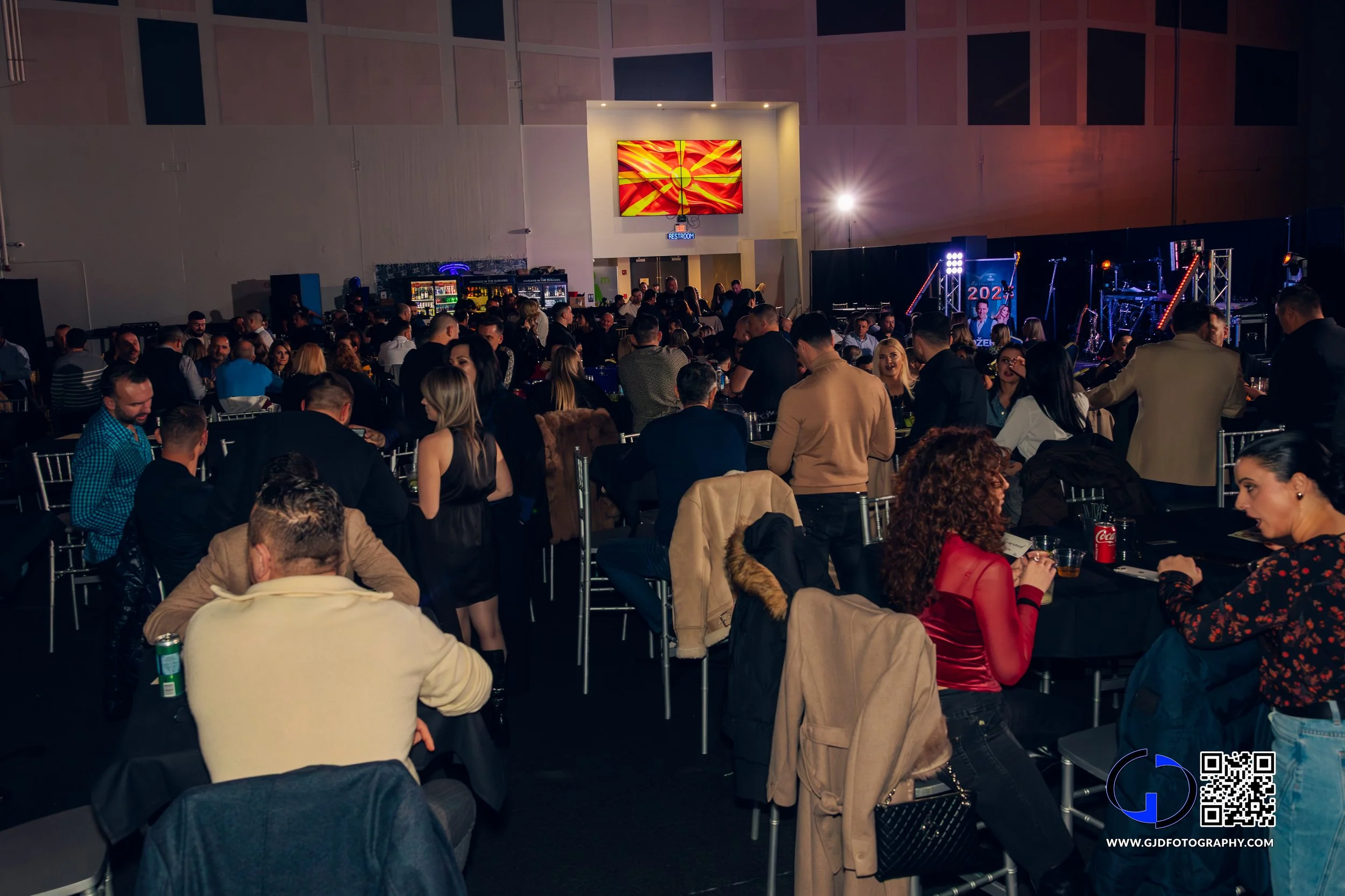 A crowded indoor event with many people sitting at round tables and some standing, with a stage that has musical equipment and lighting in the background. There is a large screen on the wall showing an abstract fiery design.