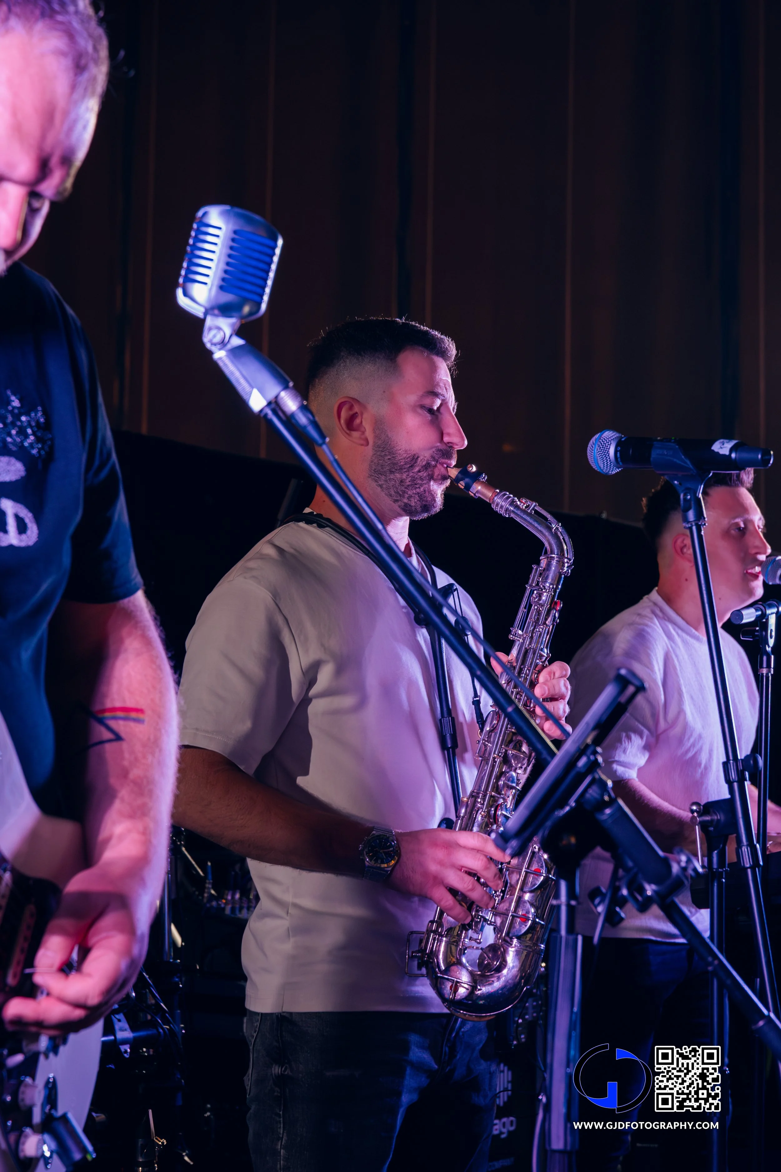 Musicians on stage, with the central focus on a bearded man playing a saxophone and singing into a microphone, while others are partially visible in the background.
