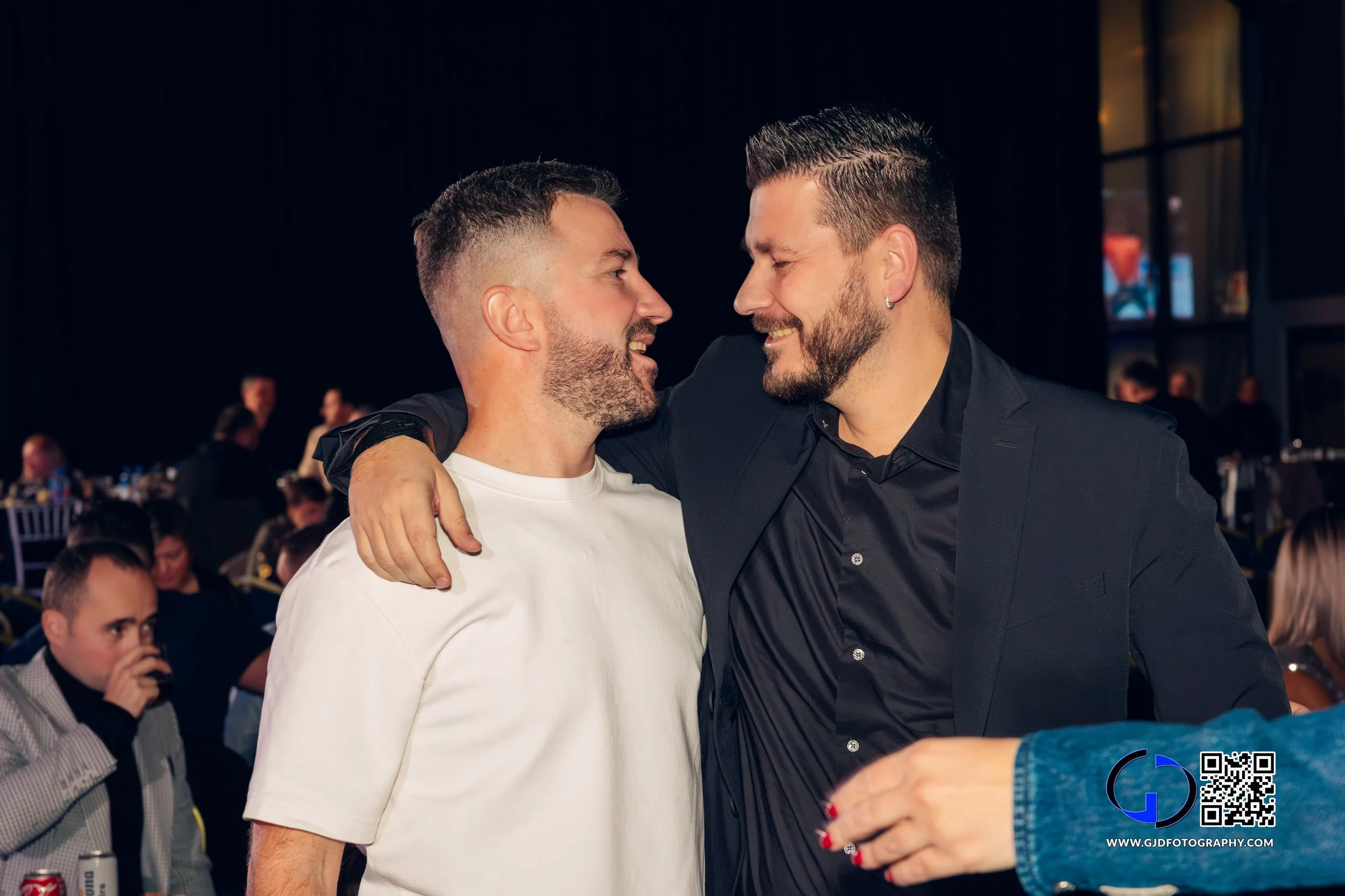 Two men with beards and short hair smiling and hugging each other at a social event, with other people sitting and standing in the background.