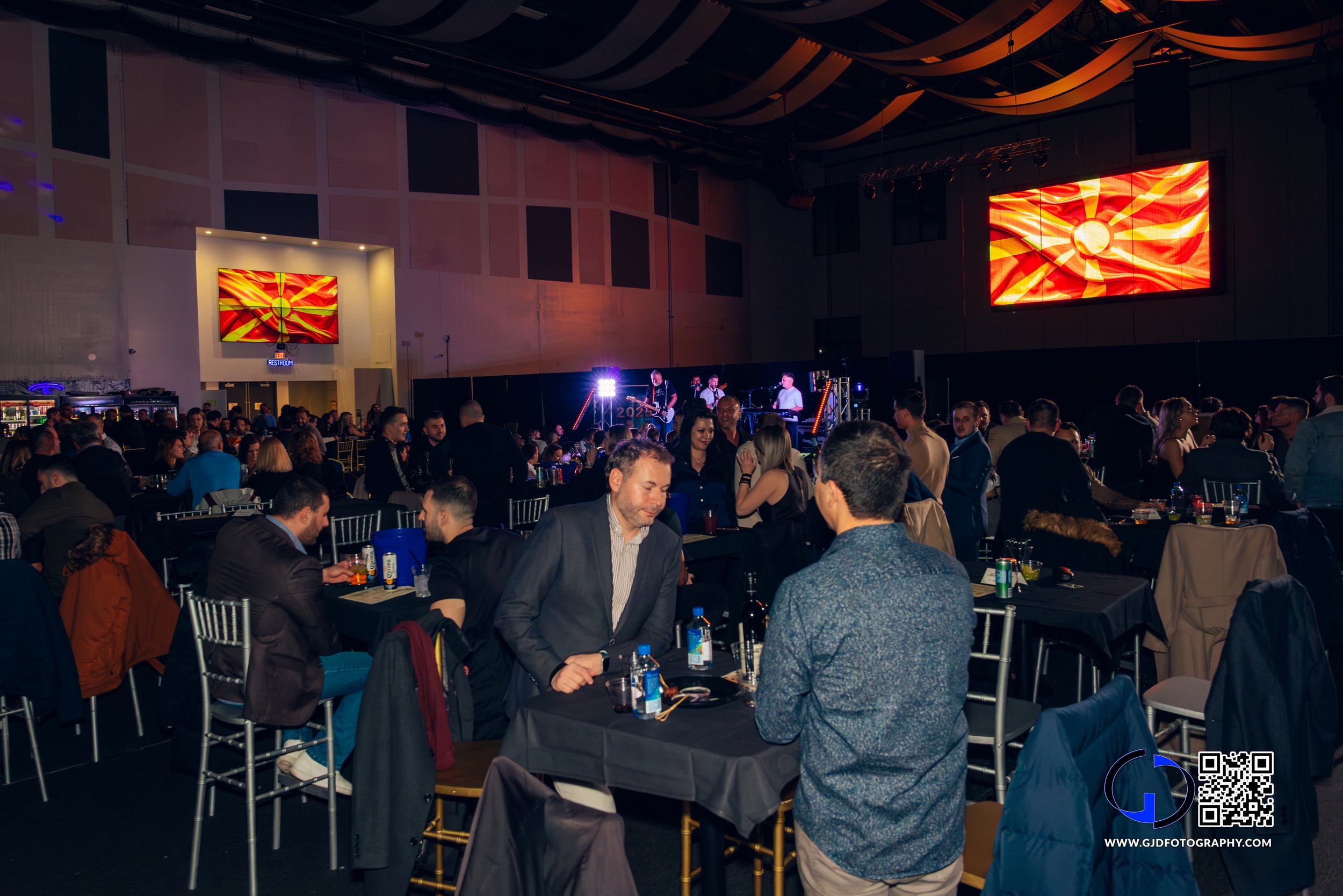 Attendees at a formal event or concert in a large indoor venue, with a stage featuring a band performing, multiple screens displaying a stylized flag or emblem, and tables with drinks and food.