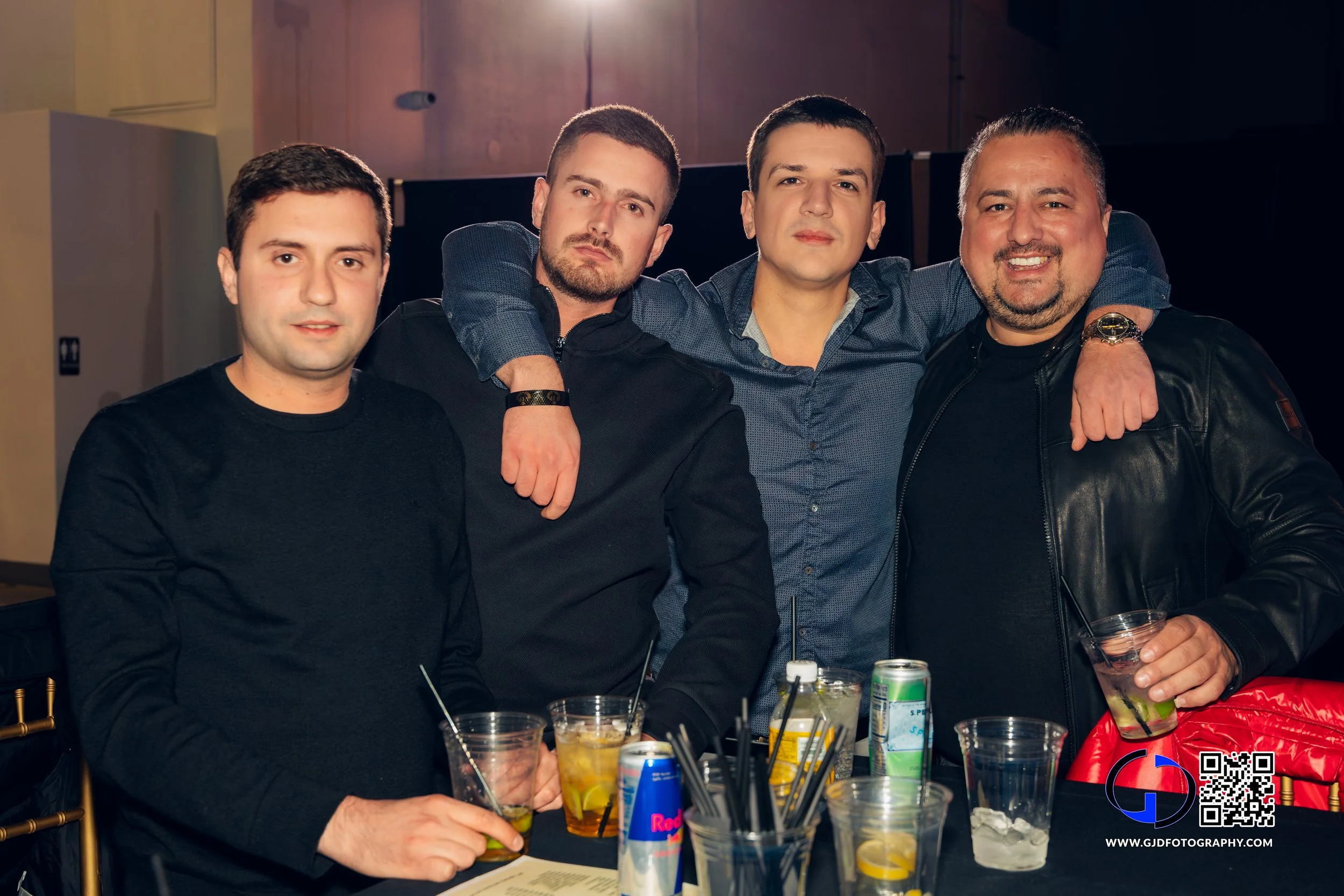 Four men standing together at a bar or restaurant, with drinks on the table in front of them. Two are wearing black, one in a blue shirt, and the fourth in a black leather jacket. They are smiling, with two men having their arms around each other's s