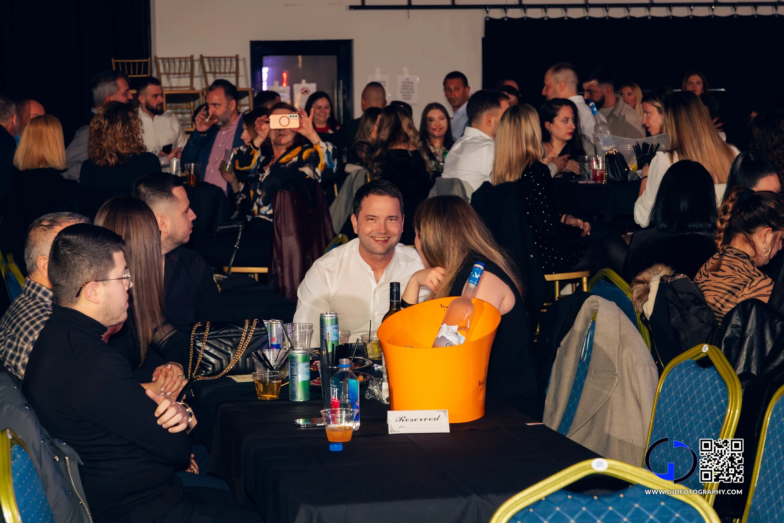 People gathered at tables in a dimly lit event hall, socializing and taking photos, with drinks and a reserved sign on the table.