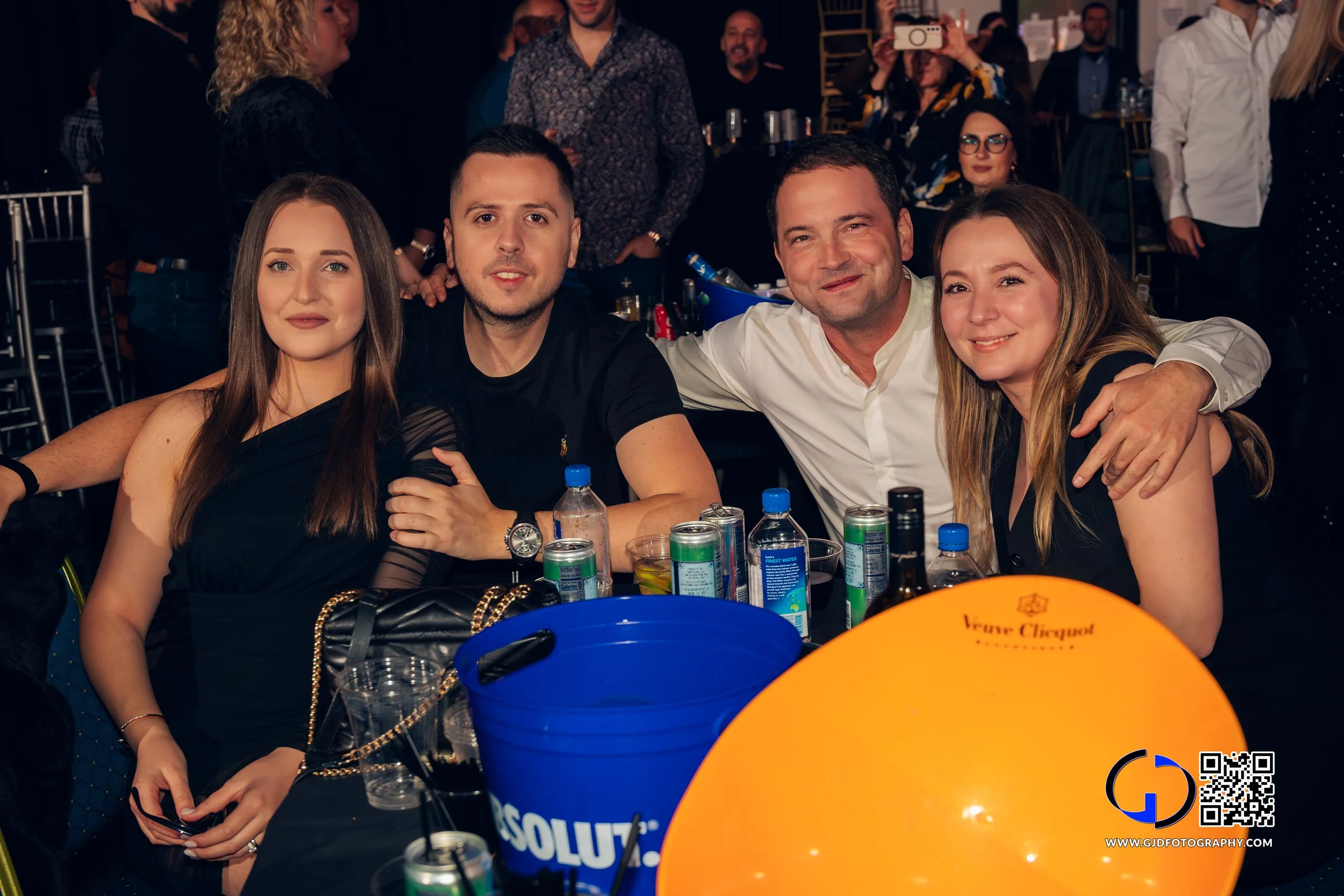 Four friends sitting at a table with drinks and cans, smiling and posing for a photo at a lively party or celebration.