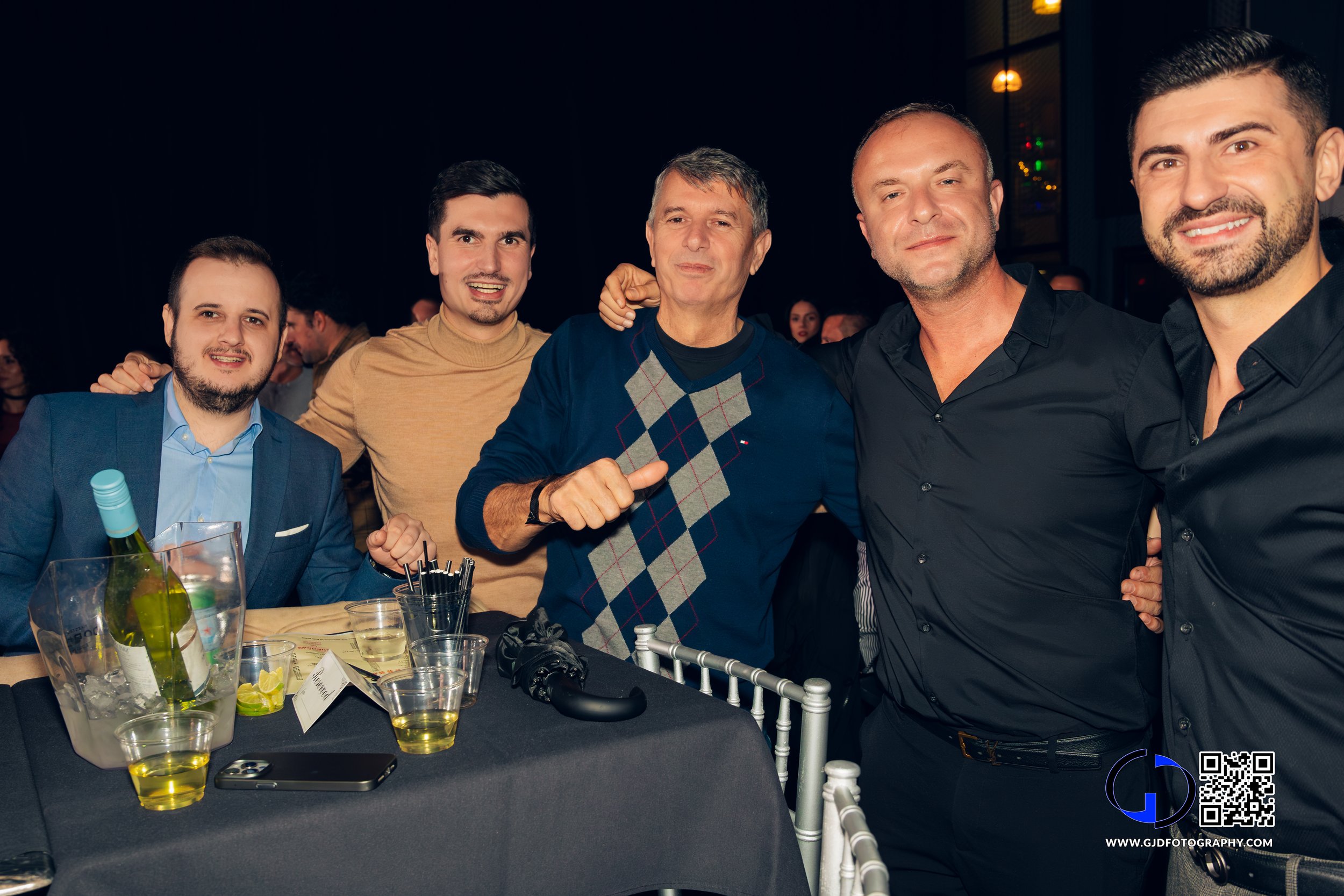 A group of five men at a social event, smiling and posing for the camera. The table in front of them has a bottle in an ice bucket, glasses of drinks, and a phone.