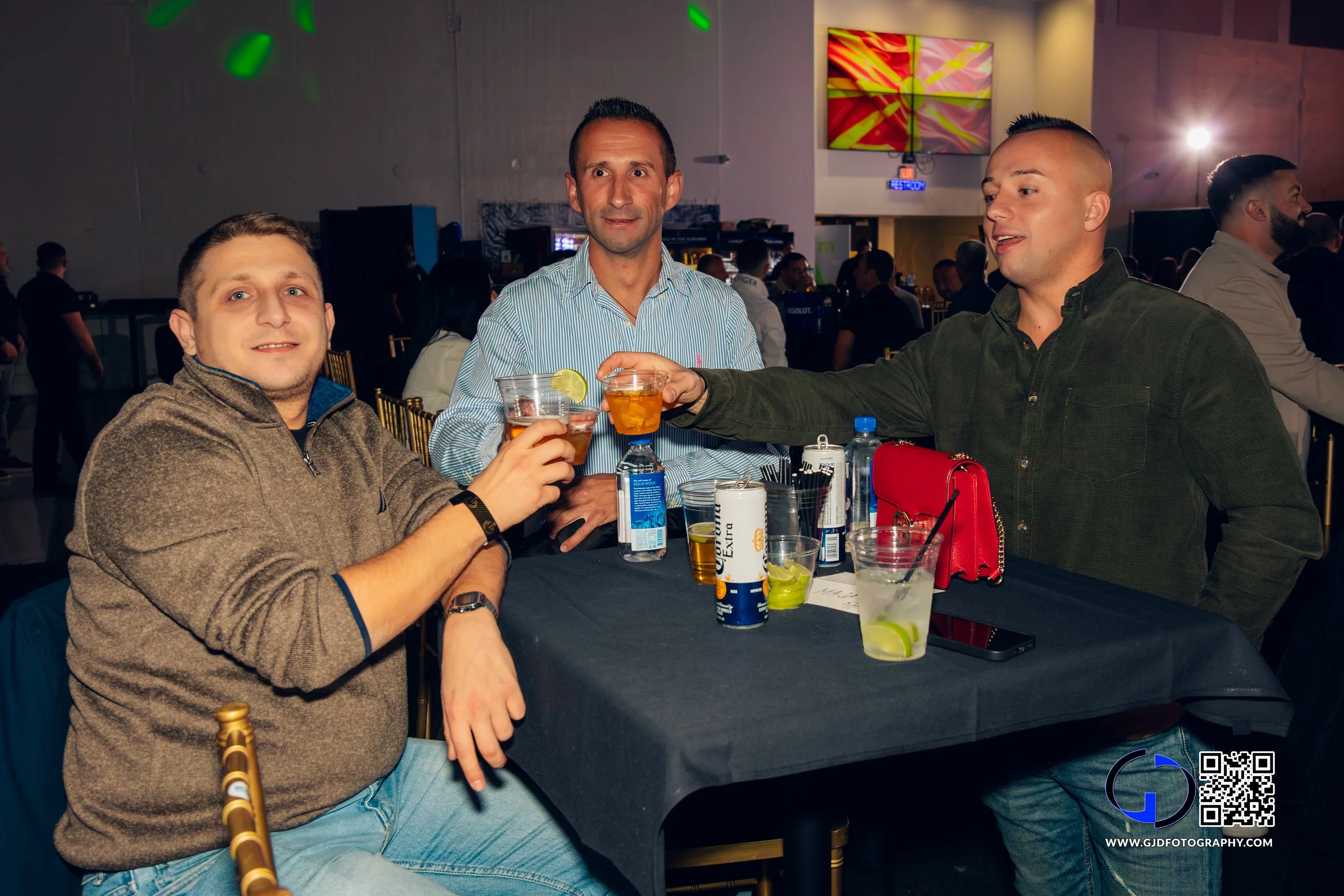 Three men sitting at a table in a bar, raising glasses for a toast, with drinks, bottles, and personal items on the table, in a lively indoor setting.