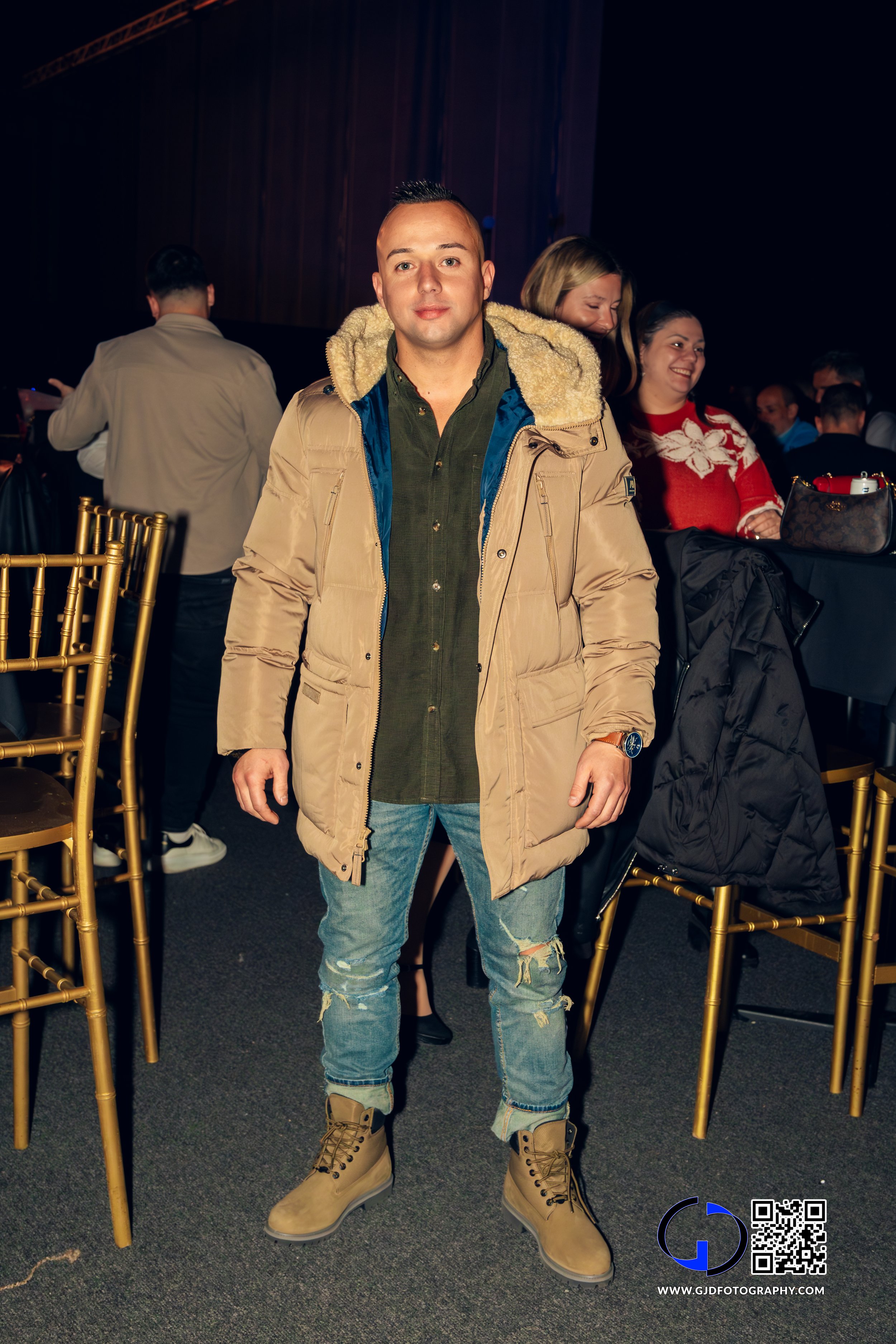 Young man standing in a crowded event space with gold chairs, wearing a beige jacket, green shirt, ripped jeans, and tan boots.
