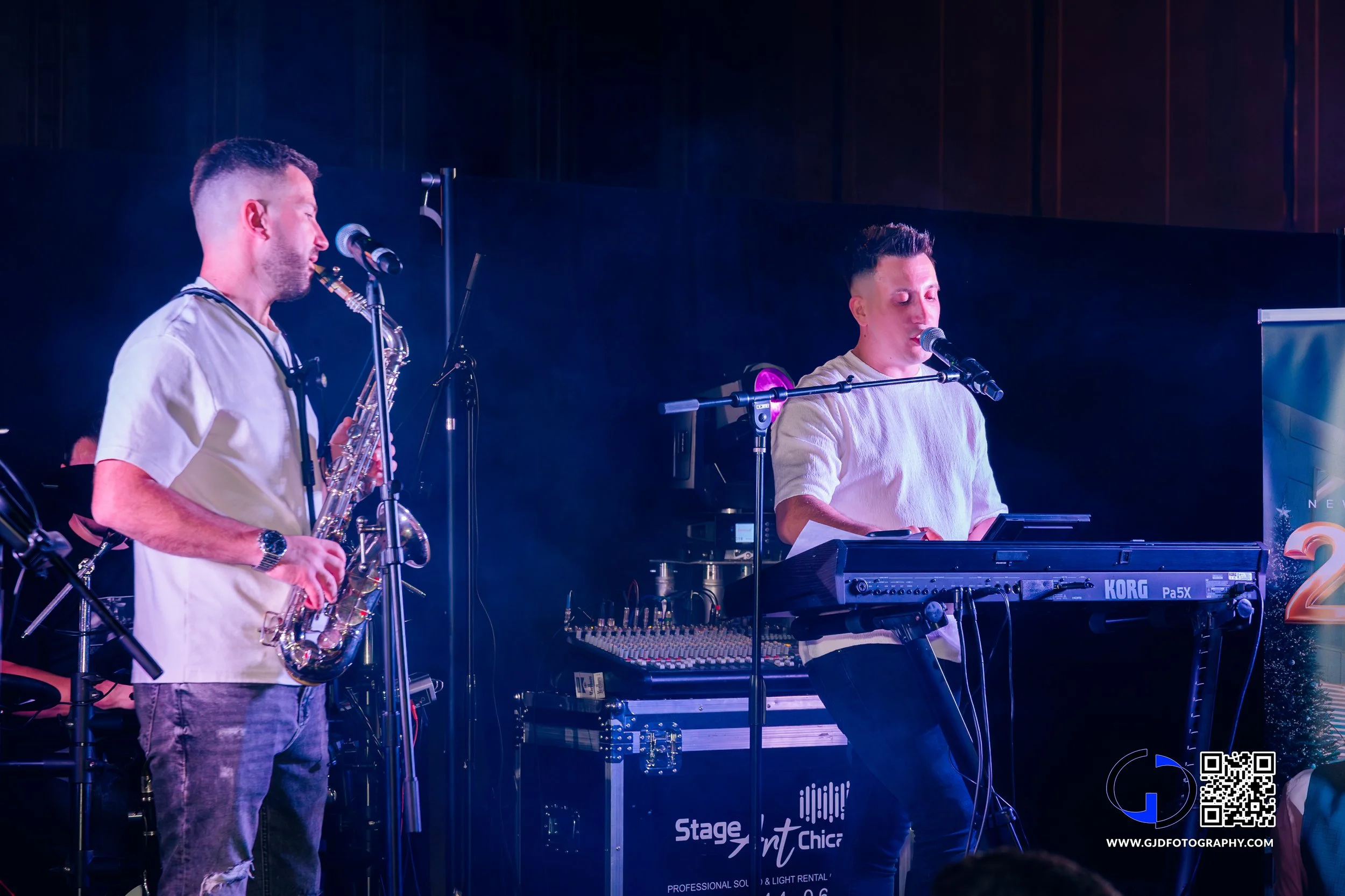 Two male musicians performing on stage, one playing a saxophone and the other singing and playing a keyboard, with microphones set up in front of them; stage equipment visible in the background.