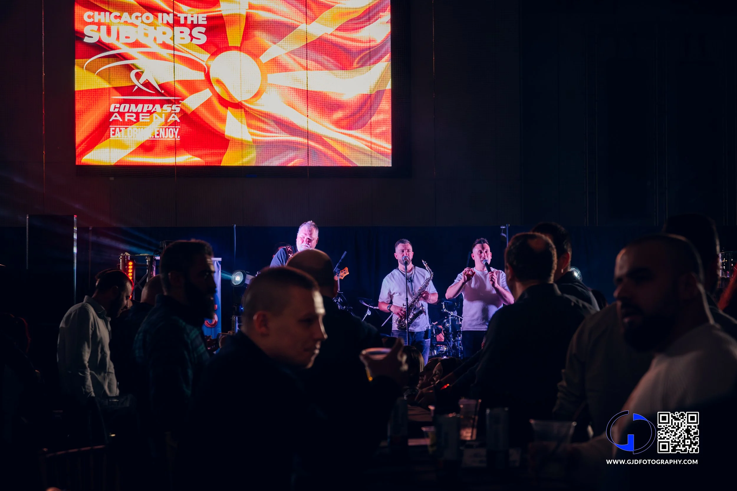 Live band performing on stage at a concert, with a large screen overhead displaying colorful graphics and text about Chicago suburbs and Compass Arena, and audience members seated and standing in front.