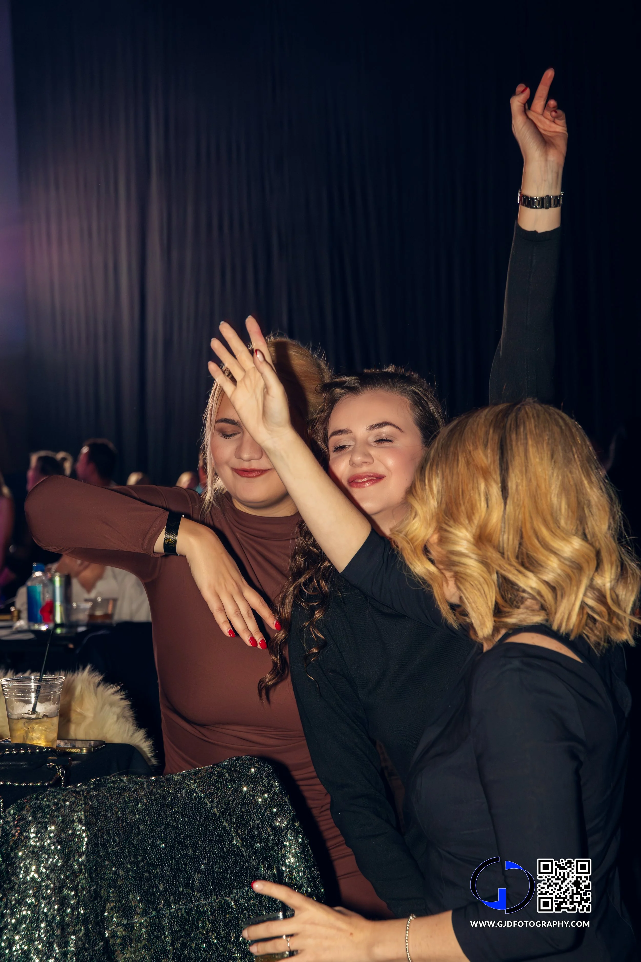 Three women dancing and enjoying themselves at a party or celebration, with others in the background and drinks on the table.